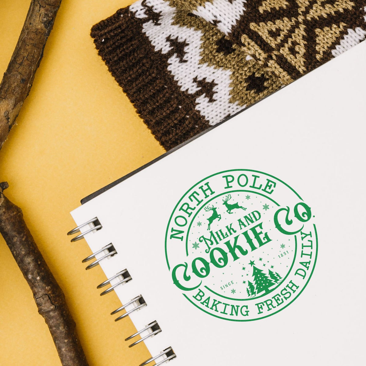 White spiral notebook with green North Pole Milk and Cookie Co. seal stamp, on yellow background with brown knit and branches.