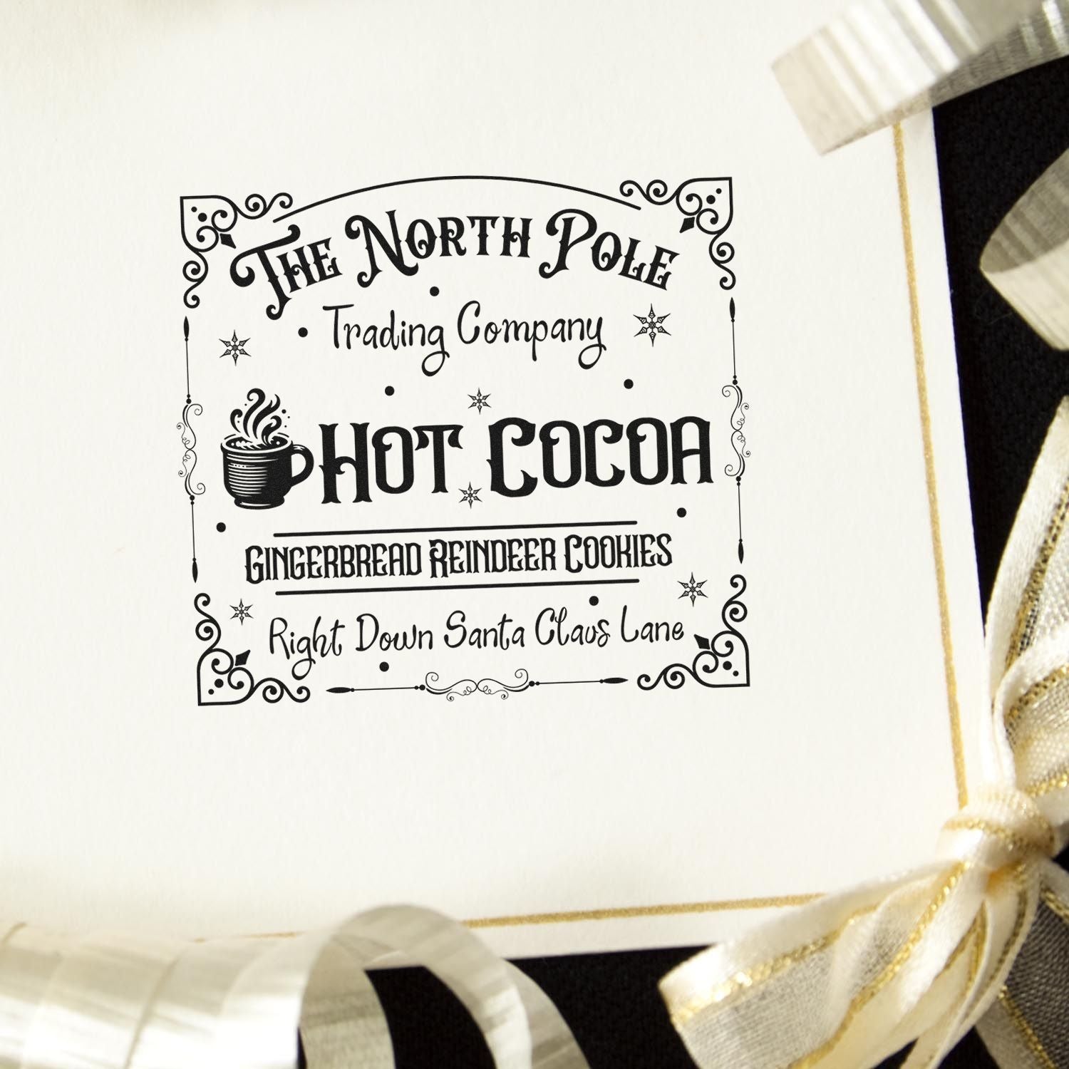 Black decorative holiday stamp seal on white card, reading The North Pole Trading Company Hot Cocoa, with cream and gold ribbon bows.