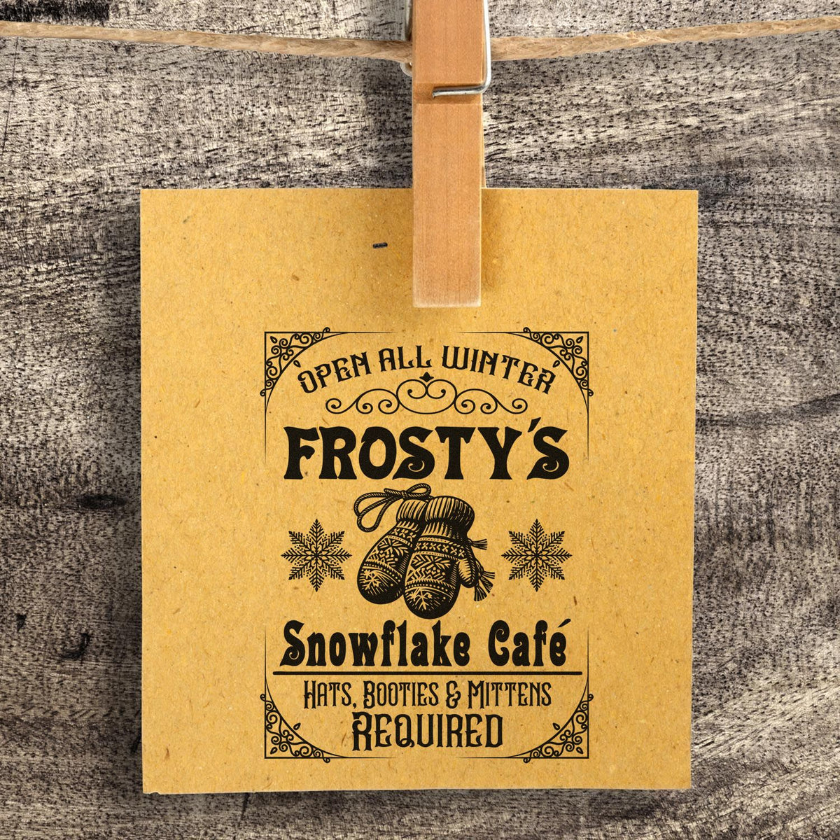 Kraft paper hanging sign with black stamp-style winter cafe design, mittens, snowflakes, and Open All Winter text.