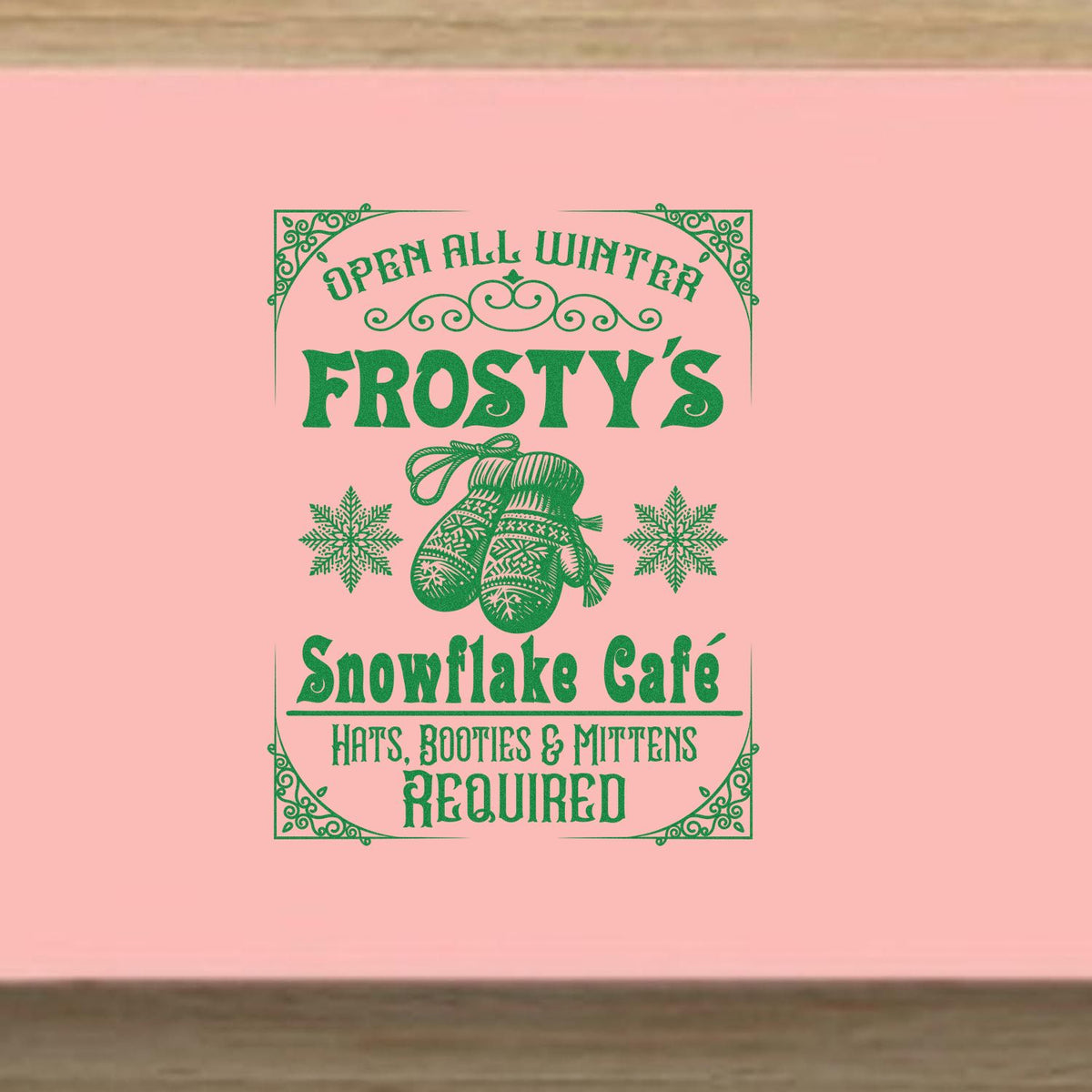 Green vintage winter stamp seal on pink background with mittens, snowflakes, and Open All Winter Frostys Snowflake Cafe text.