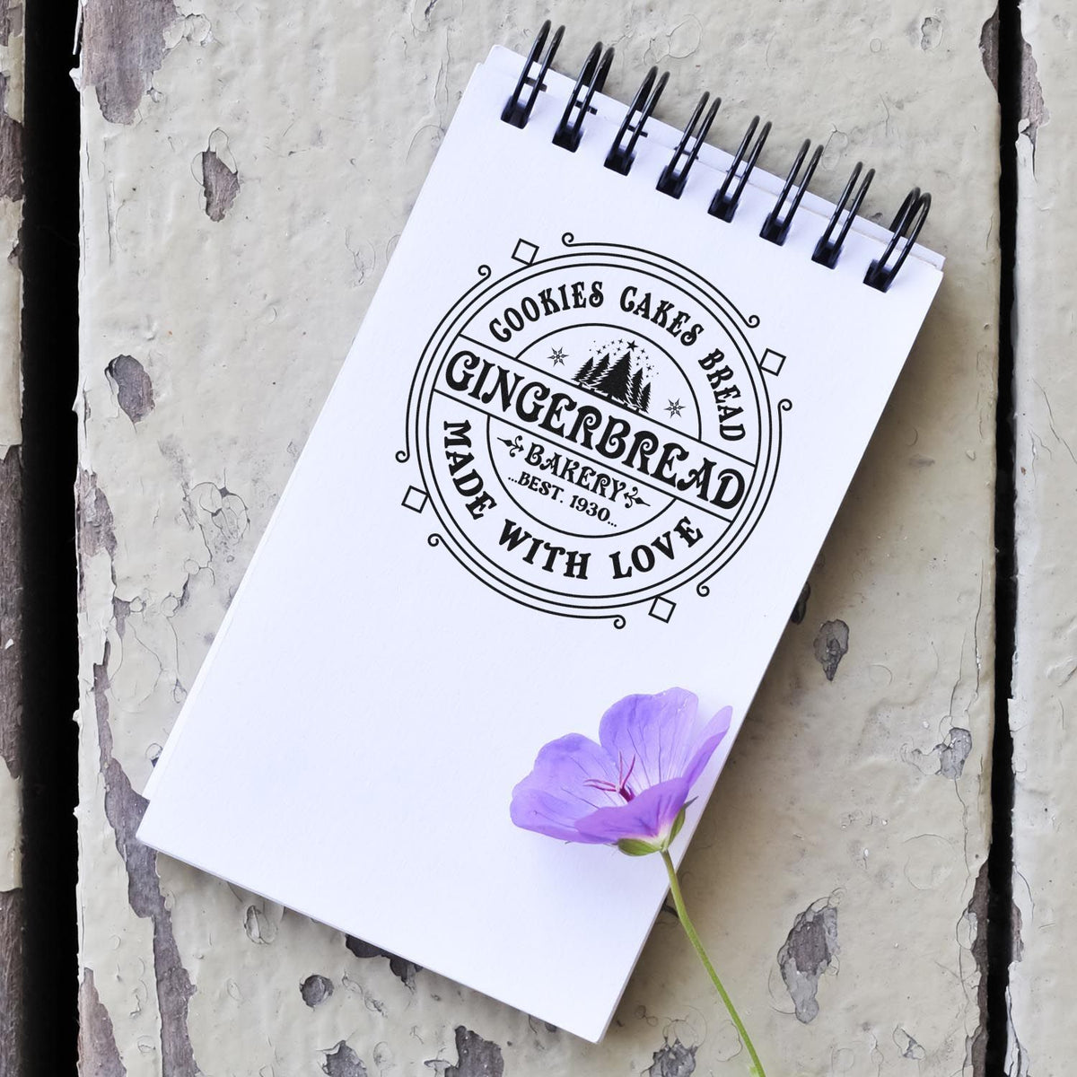 Black custom bakery stamp seal on white spiral notebook, reading Gingerbread Made With Love, with purple flower on gray wood