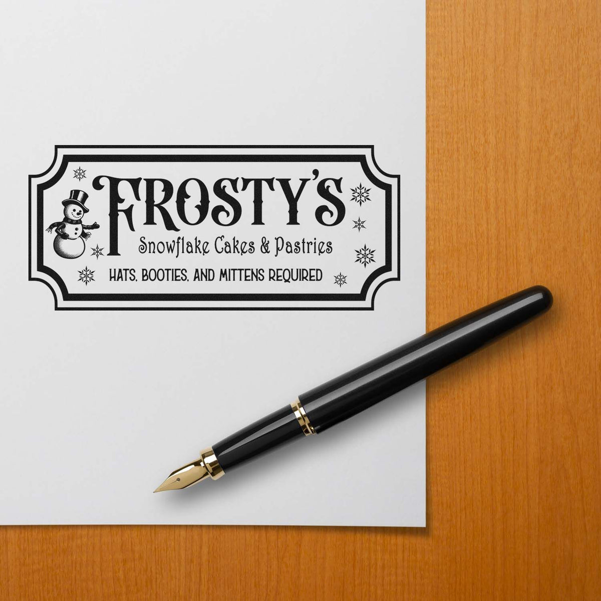 Black custom stamp seal for Frostys Snowflake Cakes & Pastries with snowman, snowflakes, and black fountain pen on white paper.