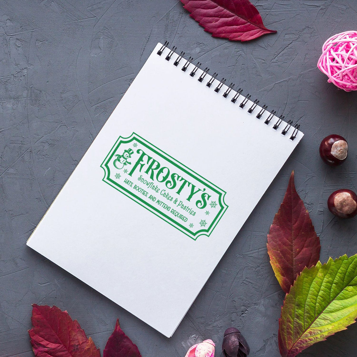 Green custom stamp seal on white spiral notebook, surrounded by autumn leaves on a gray background.