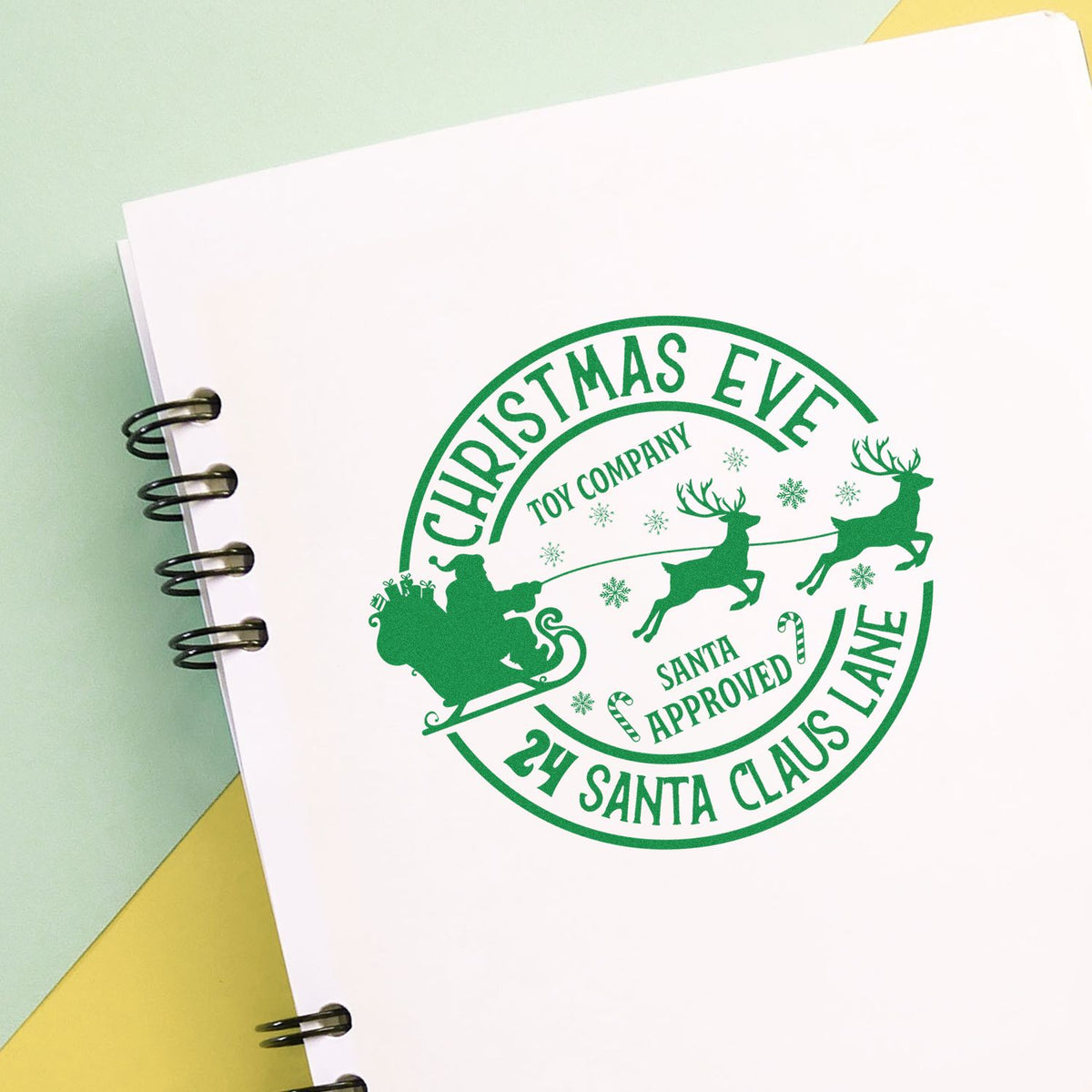 Green Christmas Eve Santa Approved circular stamp seal with sleigh, reindeer and snowflakes on a white notebook.