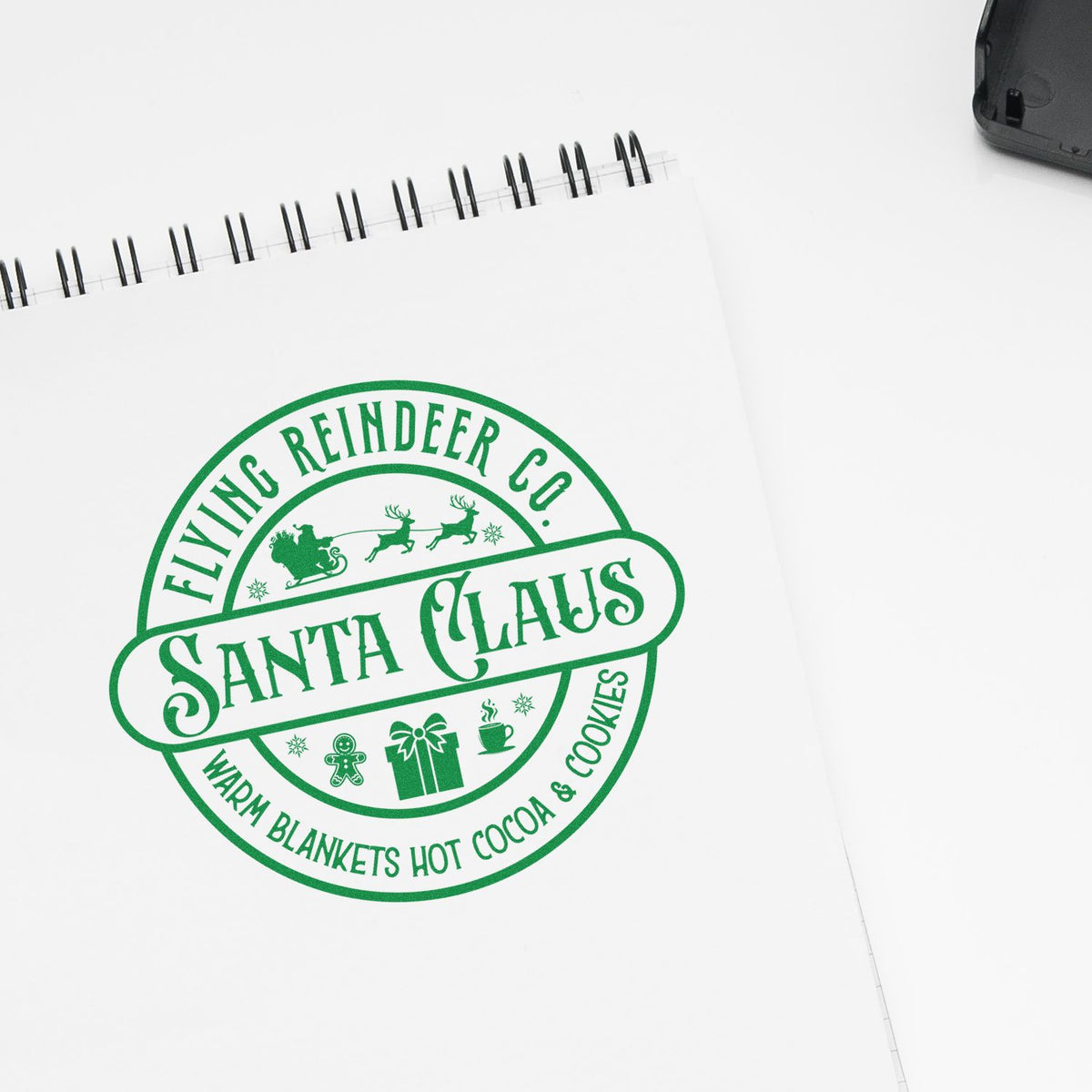 Green holiday stamp seal on white spiral notebook, reading Santa Claus with Flying Reindeer Co. and festive icons.