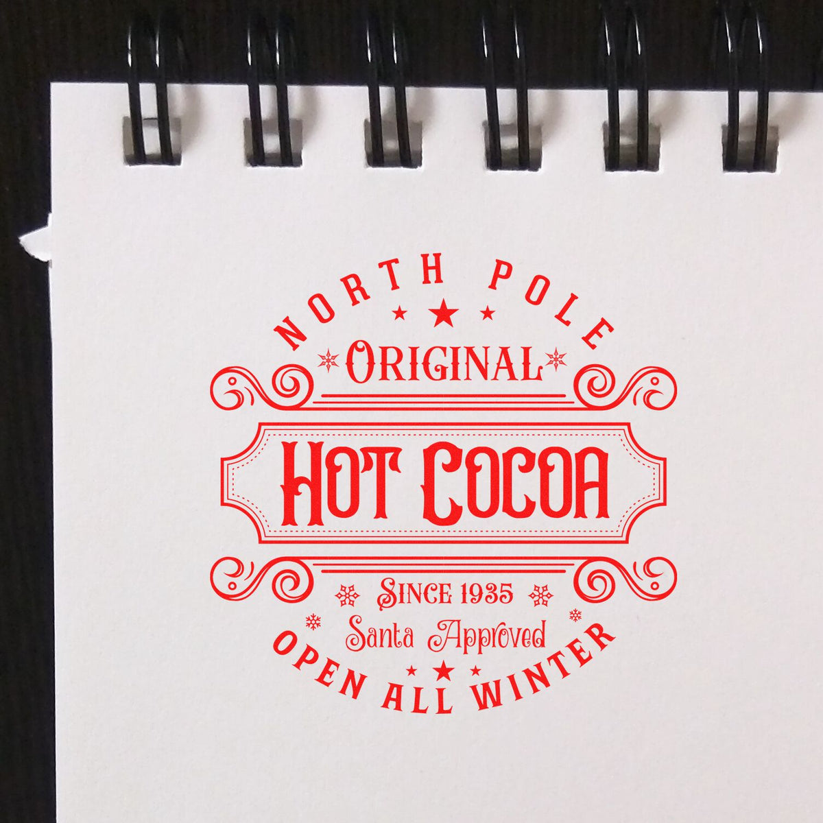 Red vintage Hot Cocoa stamp seal reading North Pole Original, Santa Approved, Open All Winter on white paper with black spiral binding.