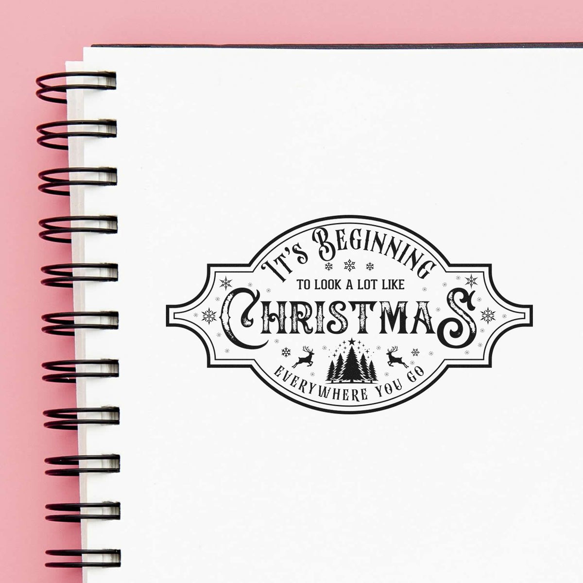 Black Christmas stamp seal on white spiral notebook page with pink background and festive trees, reindeer, and snowflakes.