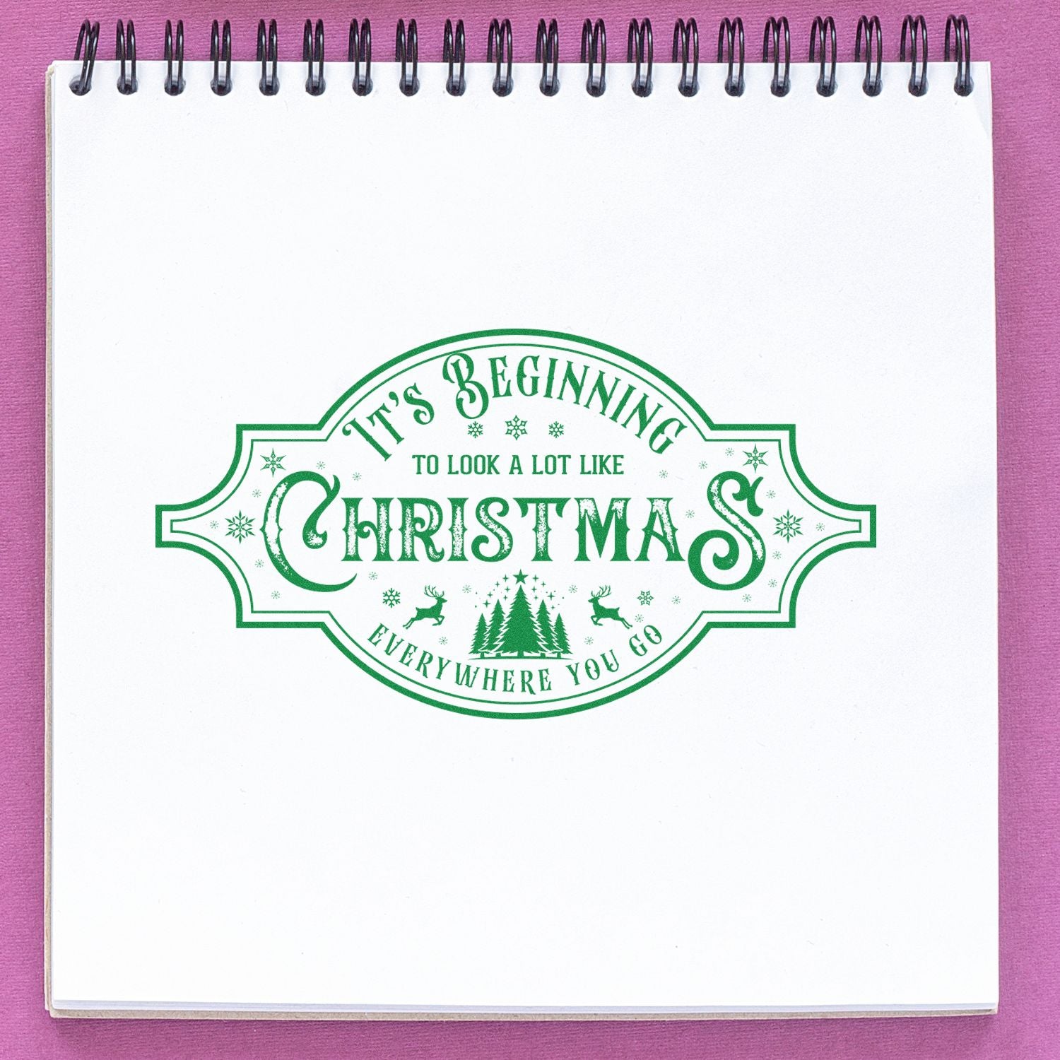 Green Christmas stamp seal reading Its Beginning to Look a Lot Like Christmas on a white spiral notebook with pink background.