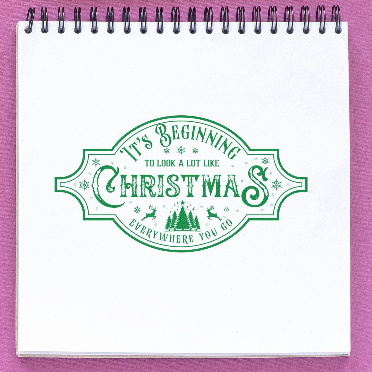 Green Christmas stamp and seal design on white spiral notebook, with trees, snowflakes, and reindeer on a pink background.