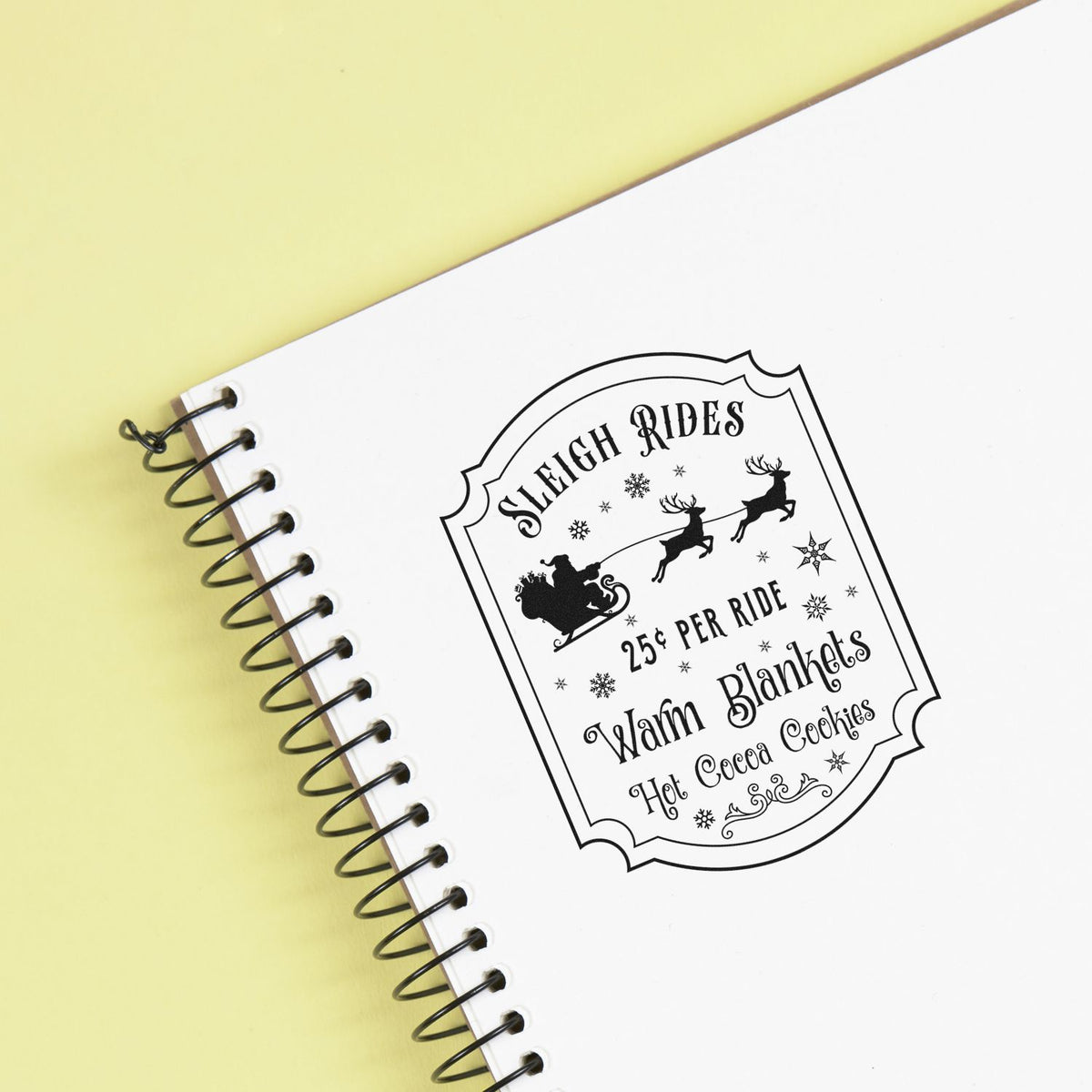 Black Sleigh Rides stamp seal on white spiral notebook, with Santa sleigh, reindeer and snowflakes on pale yellow background.