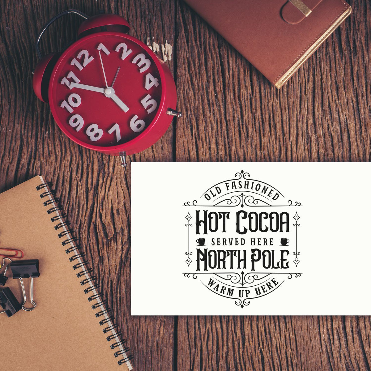 Vintage black hot cocoa stamp and seal design on white card, beside a red alarm clock on rustic wood.