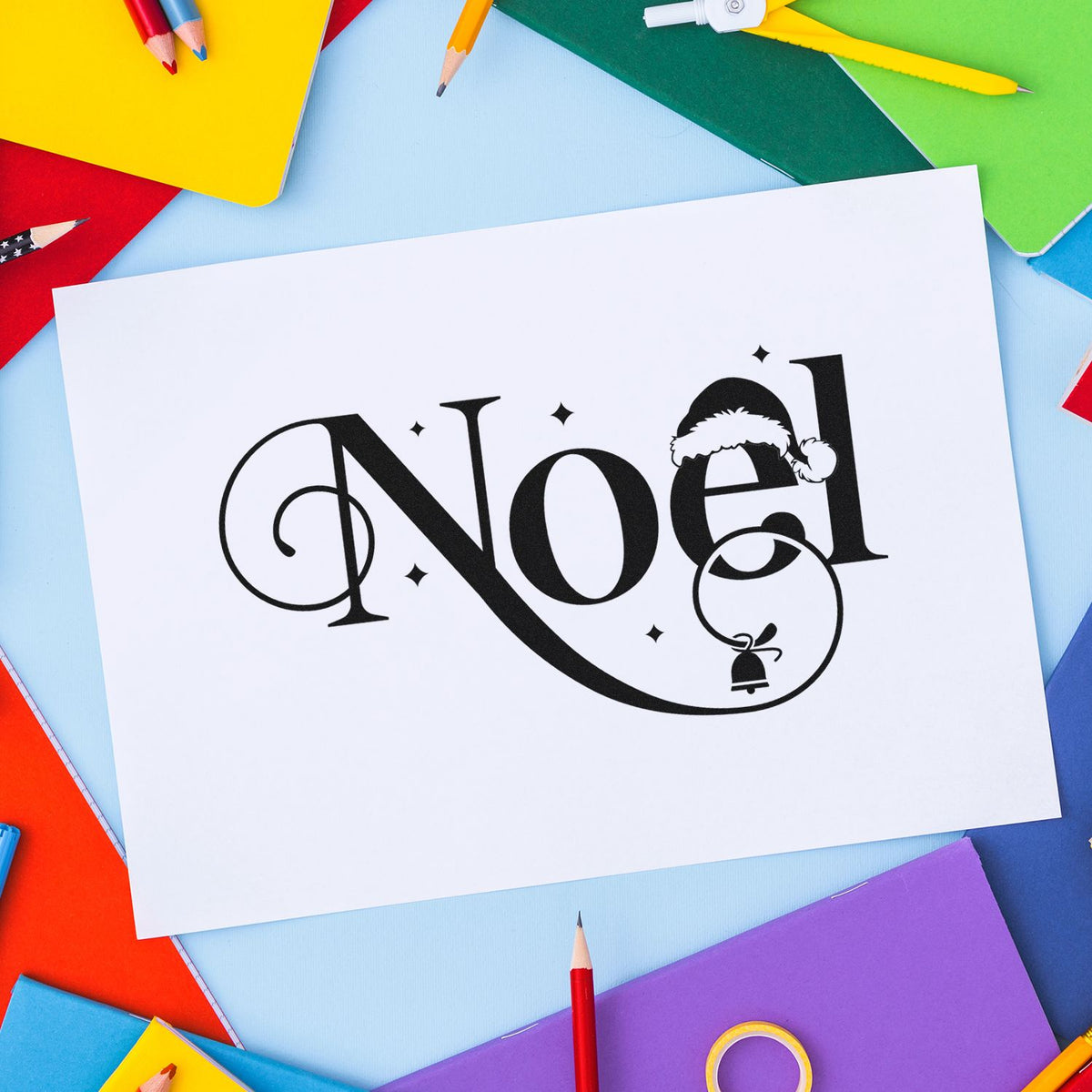 Black Noel holiday stamp design on white paper, with Santa hat and bell, surrounded by colorful notebooks and pencils.