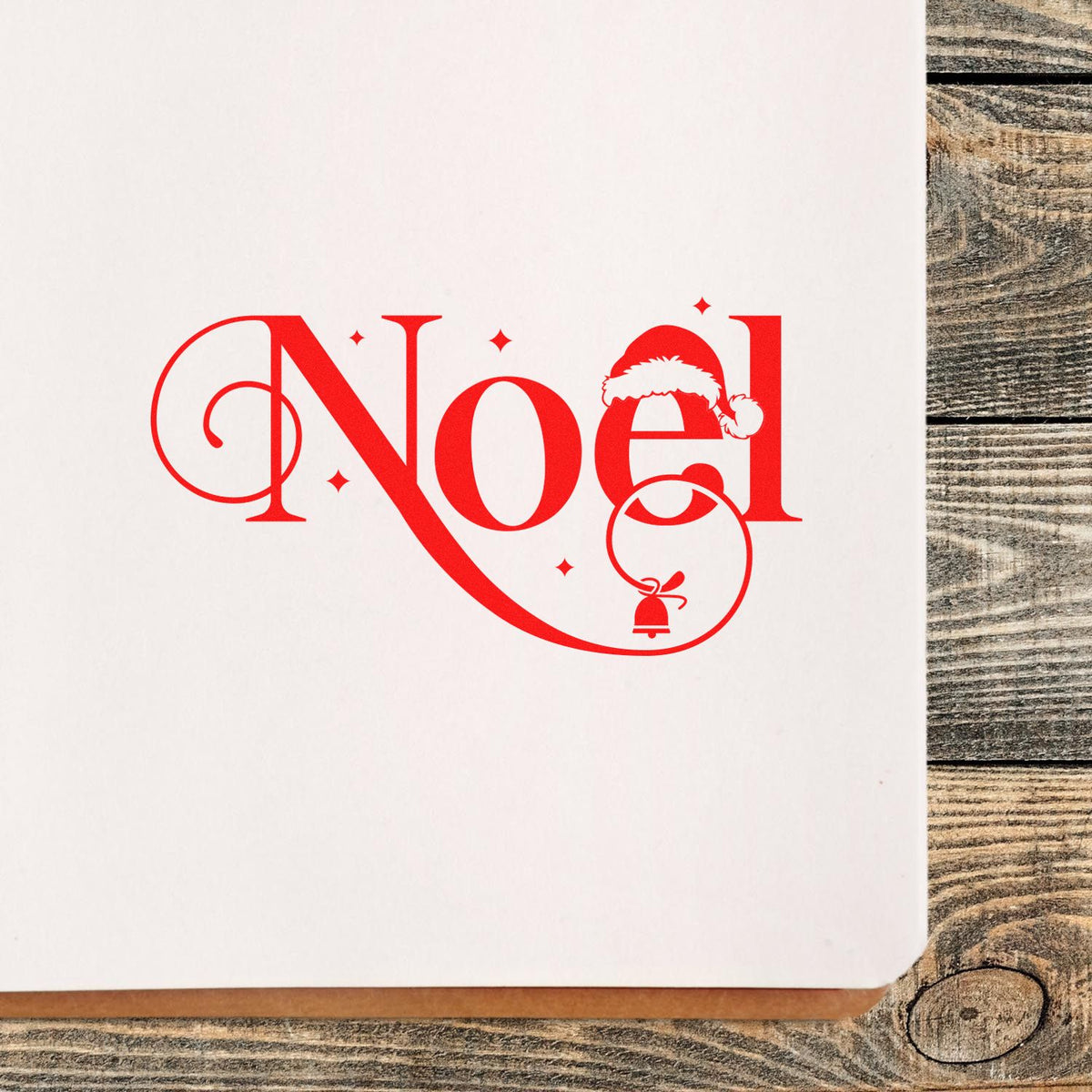Red Noel holiday stamp and seal on white card, with Santa hat, bell, and stars on rustic wood background.