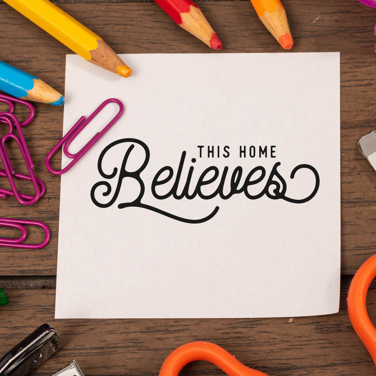 Black "This Home Believes" stamp/seal on white paper, framed by colorful pencils, purple paper clips, and wood desk.