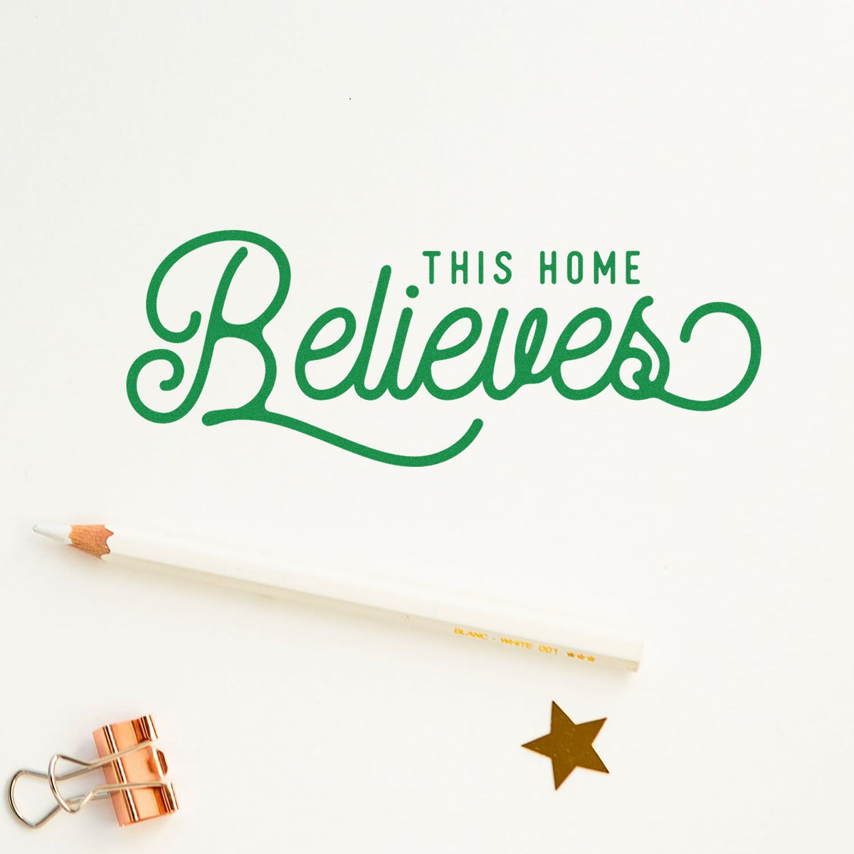 Green This Home Believes stamp-style lettering on white, with pencil, copper binder clip, and gold star seal accent.