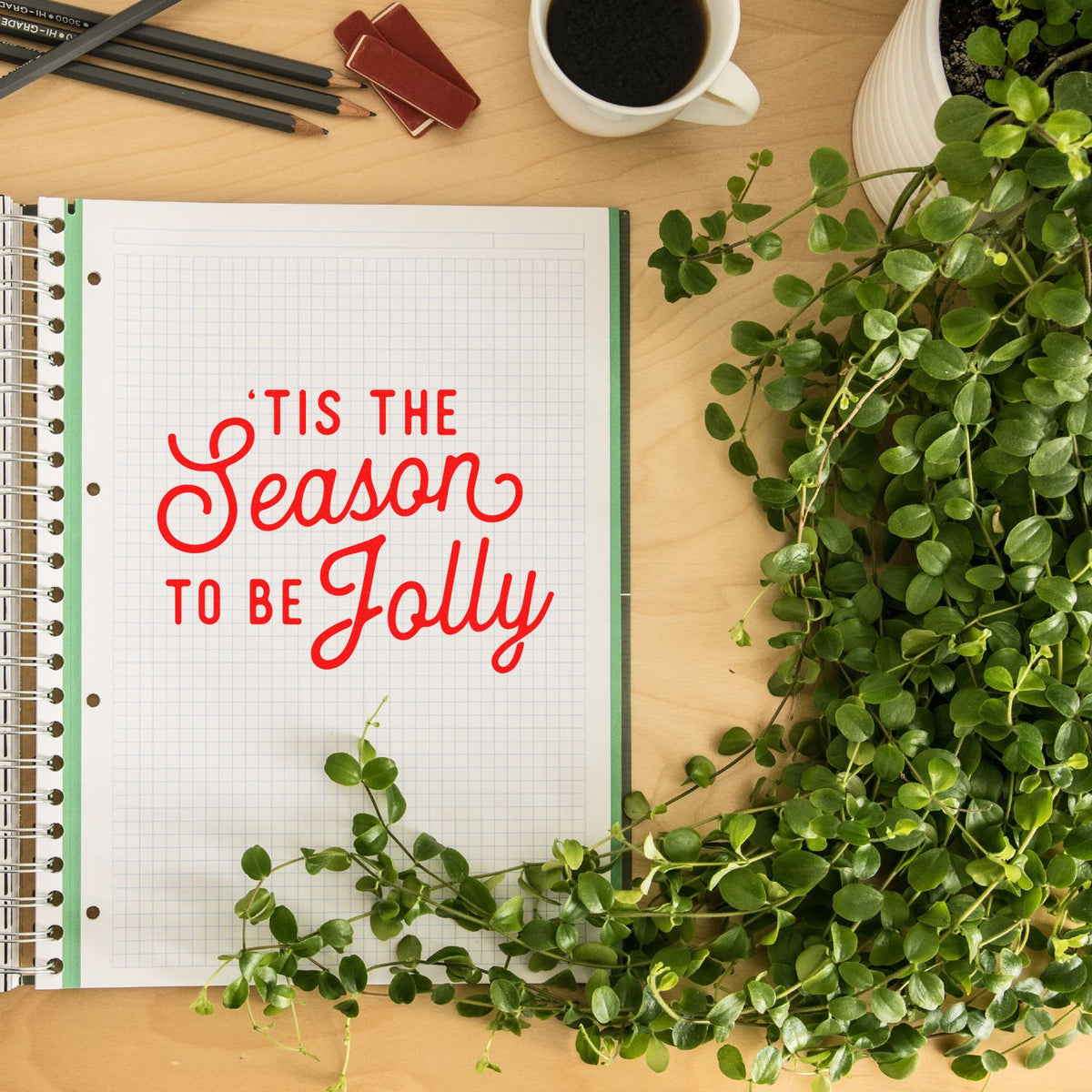 Red holiday stamp and seal reading Tis the Season to Be Jolly on graph paper, with green leaves and desk items.
