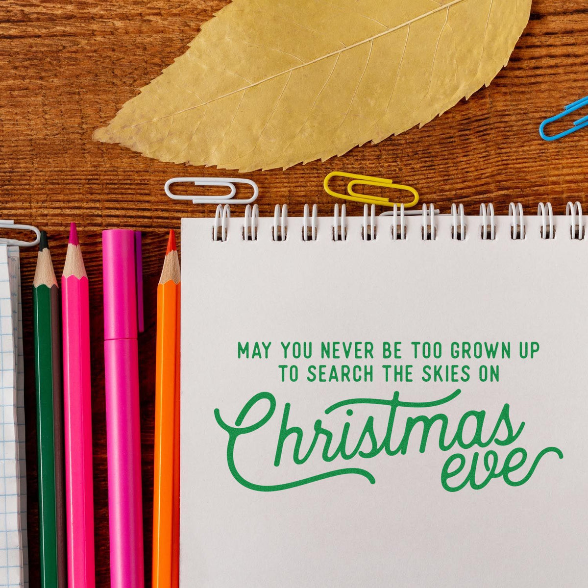 Christmas stamp and seal design on spiral notebook, with green lettering, colorful pencils, paper clips, and a tan leaf on wood.