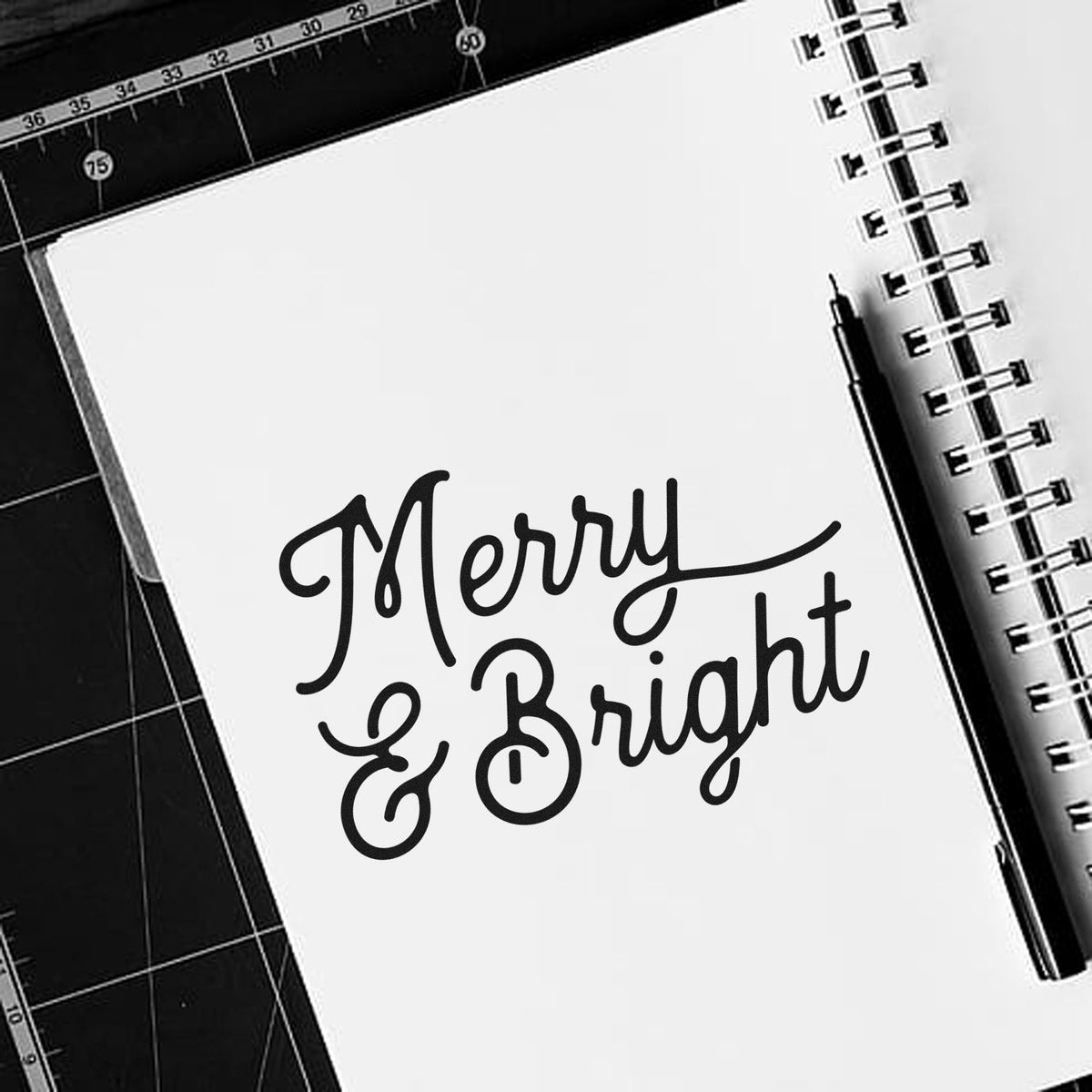 Merry & Bright black script stamp on a white spiral notebook, with black pen on a black cutting mat