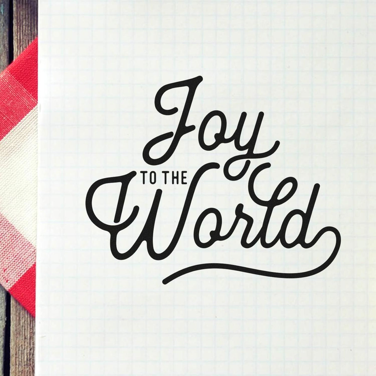 Black Joy to the World script stamp and seal on white grid paper, with red plaid and wood accents.