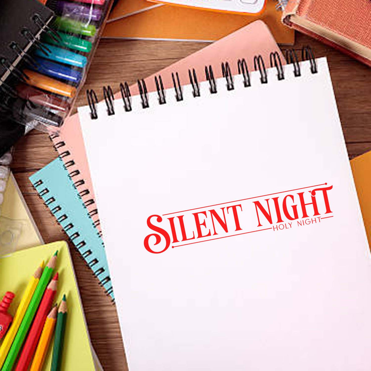 Red Silent Night Holy Night stamp seal on white spiral notebook, with colorful notebooks and pencils.