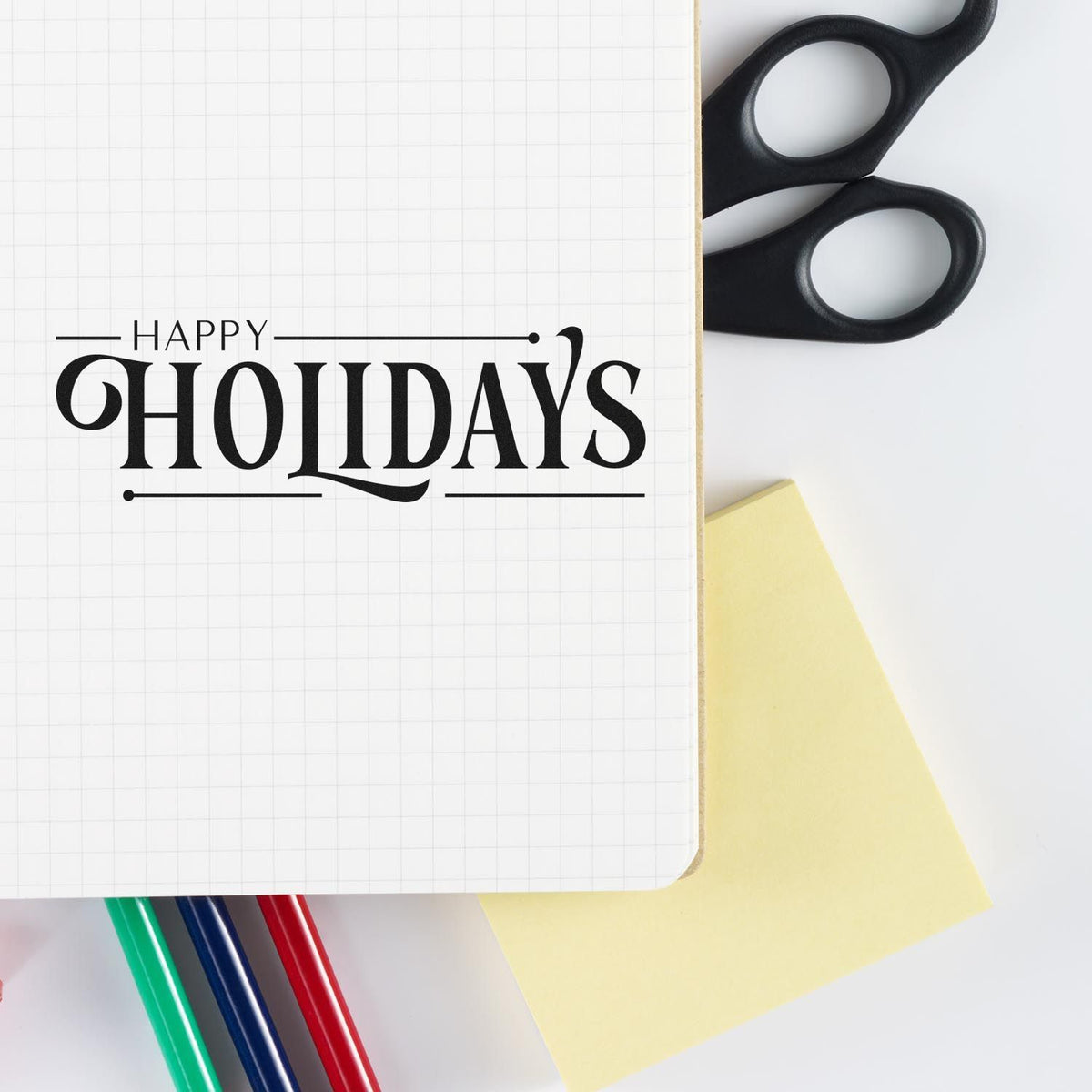 Black Happy Holidays stamp on grid notebook with black scissors, yellow sticky note, and green, blue, red pens.