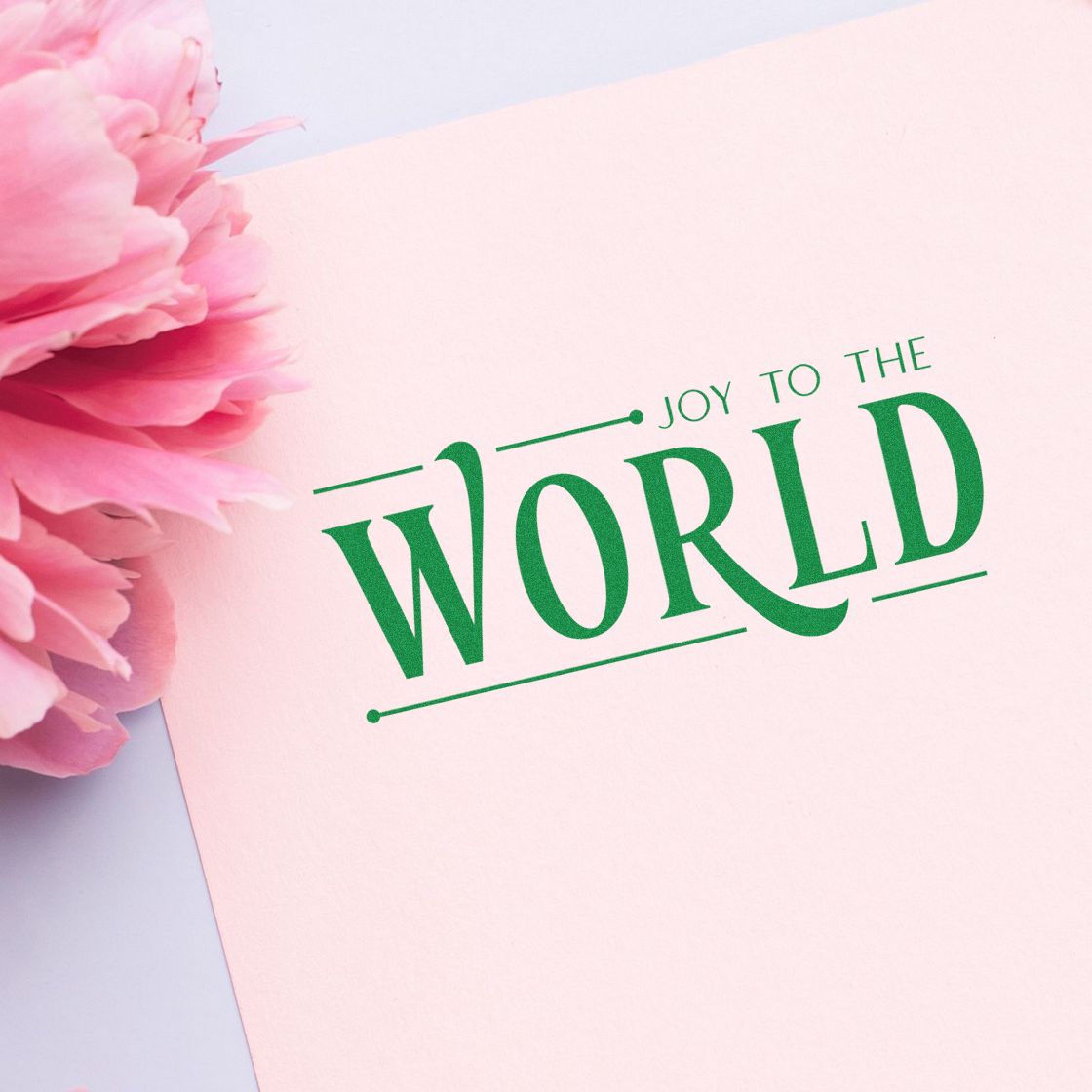 Green holiday stamp seal reading JOY TO THE WORLD on pale pink card with pink peony petals.