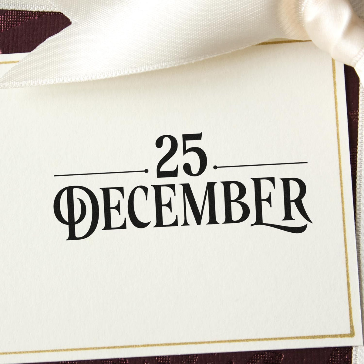 Cream stamp and seal card with black 25 December text, gold border, and satin ribbon on a burgundy background.