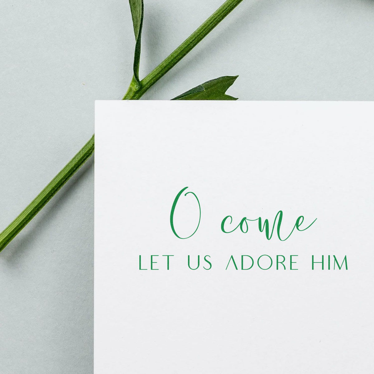 Green stamped O come let us adore him on a white card with leafy stem accents for stamp and seal branding.