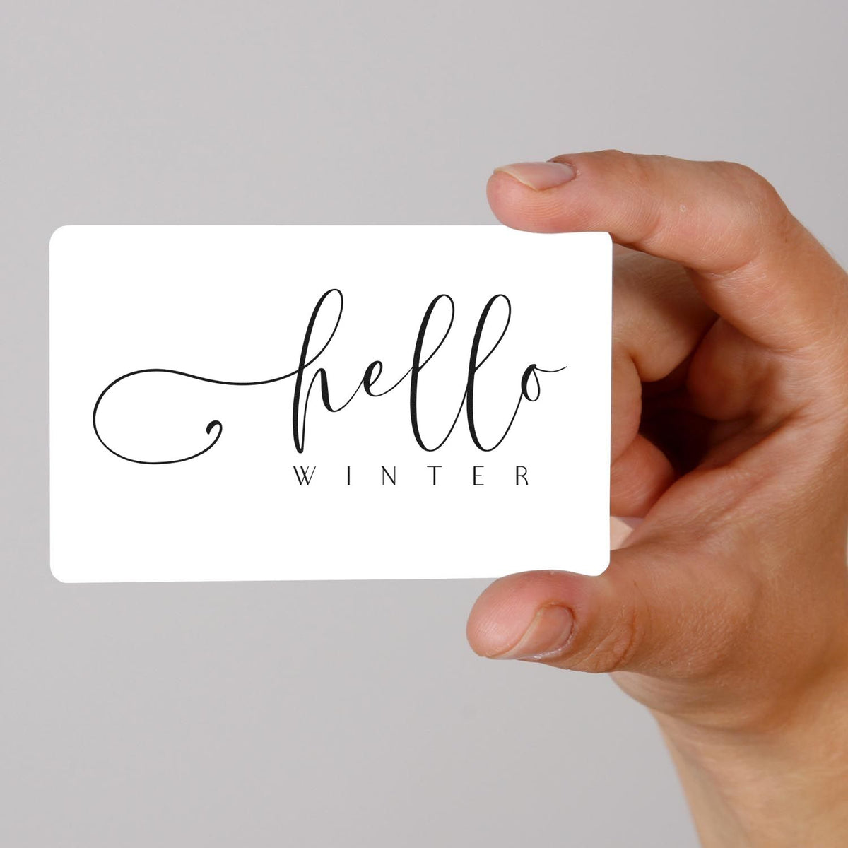 Hand holding white stamp and seal card with black hello winter script on a gray background.