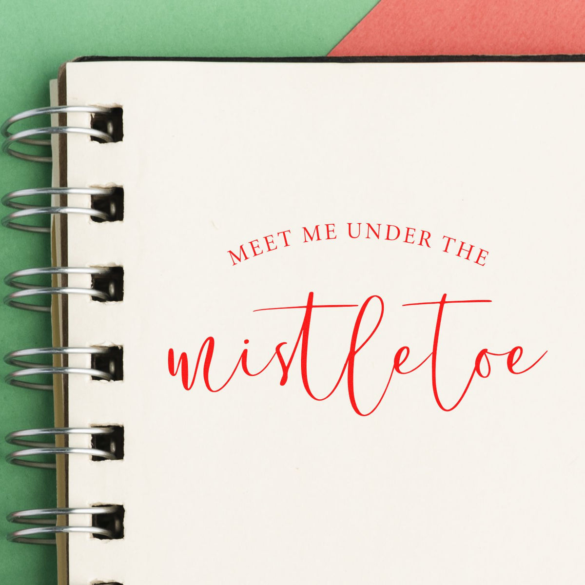 Red mistletoe holiday stamp on cream spiral-bound notebook page, with green and red background accents.