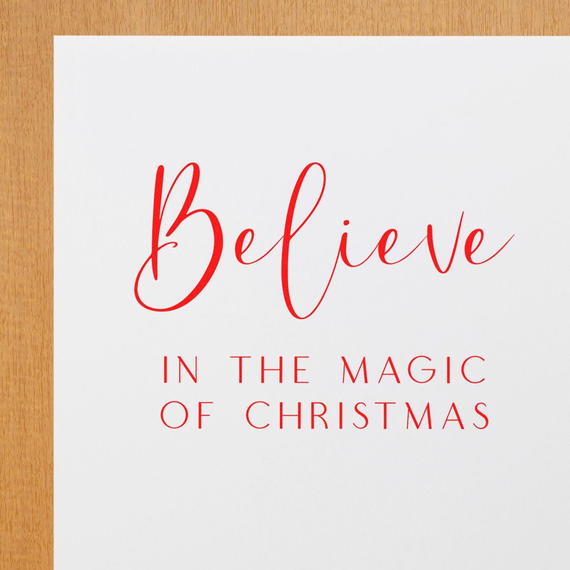 Red Christmas stamp and seal on white paper reading Believe in the Magic of Christmas on a wood surface.