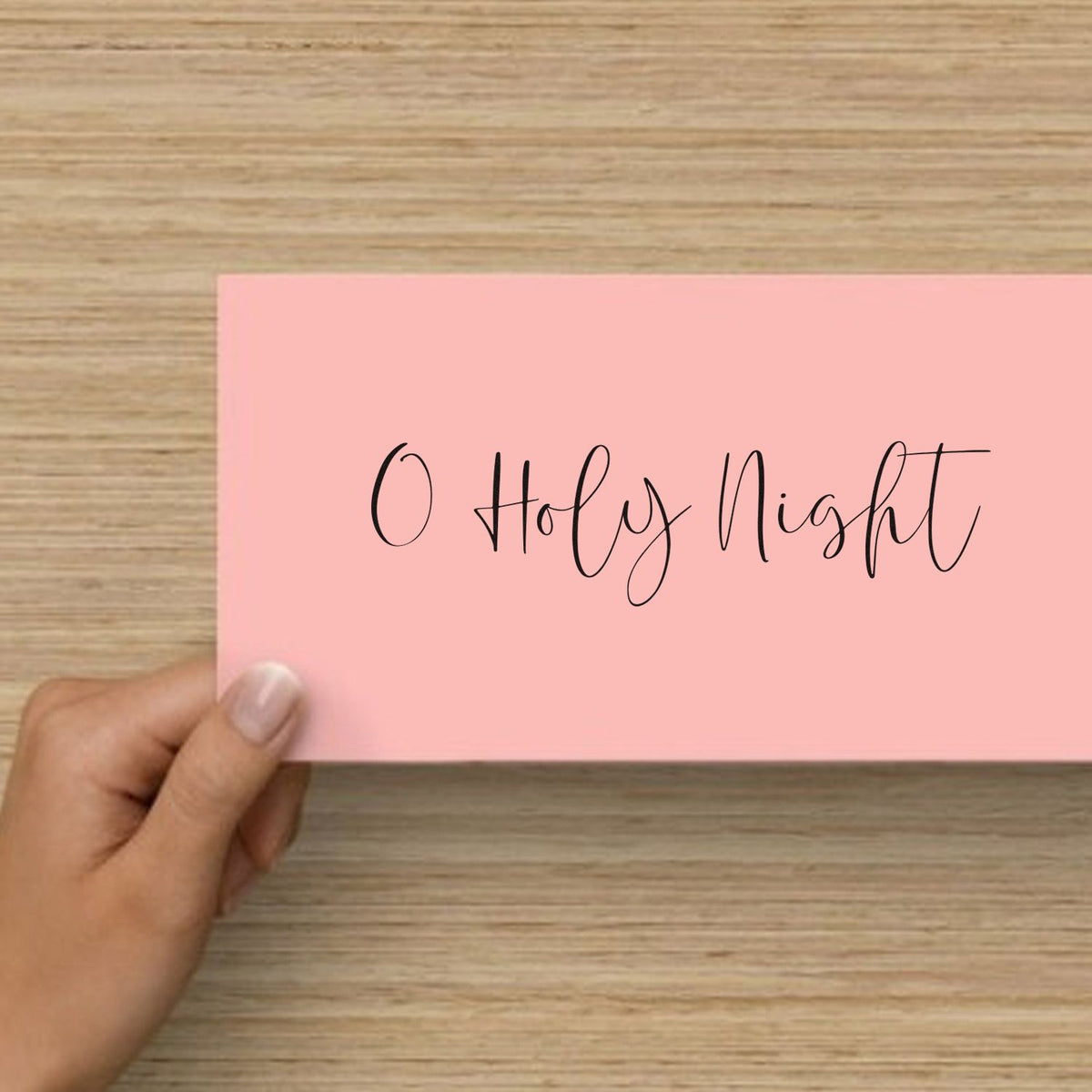 Pink stamp seal card with black O Holy Night script, held by hand over a wood tabletop.