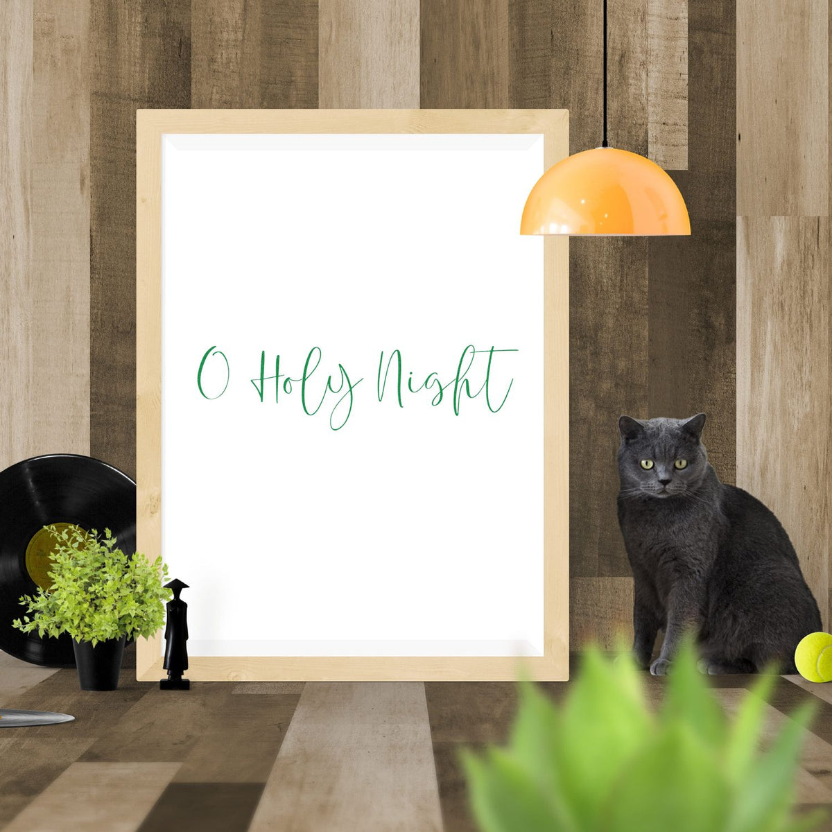 Green O Holy Night holiday stamp and seal mockup on white framed board with wood backdrop, orange lamp, gray cat and plants