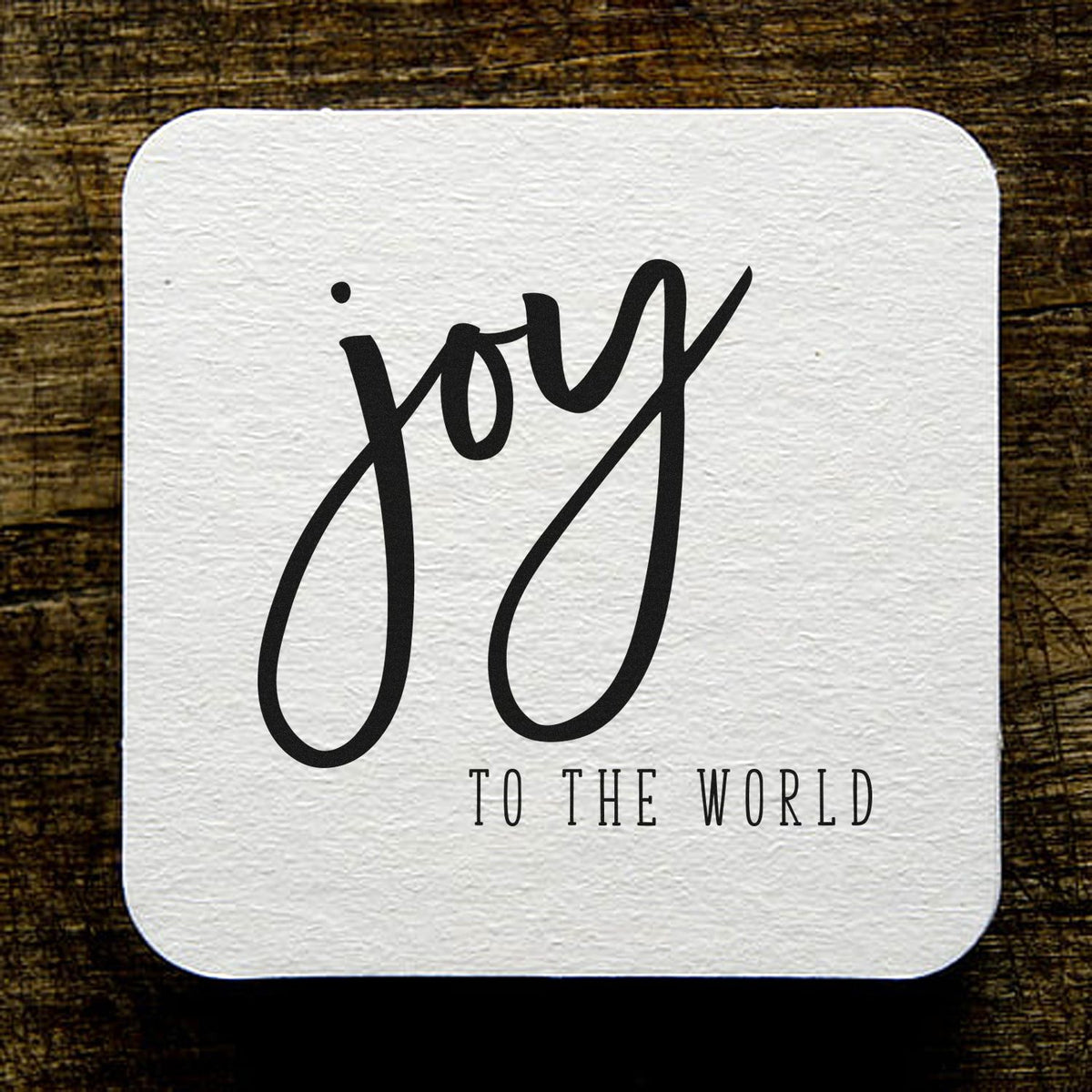 White textured square stamp/seal with black joy to the world lettering on rustic wood background.