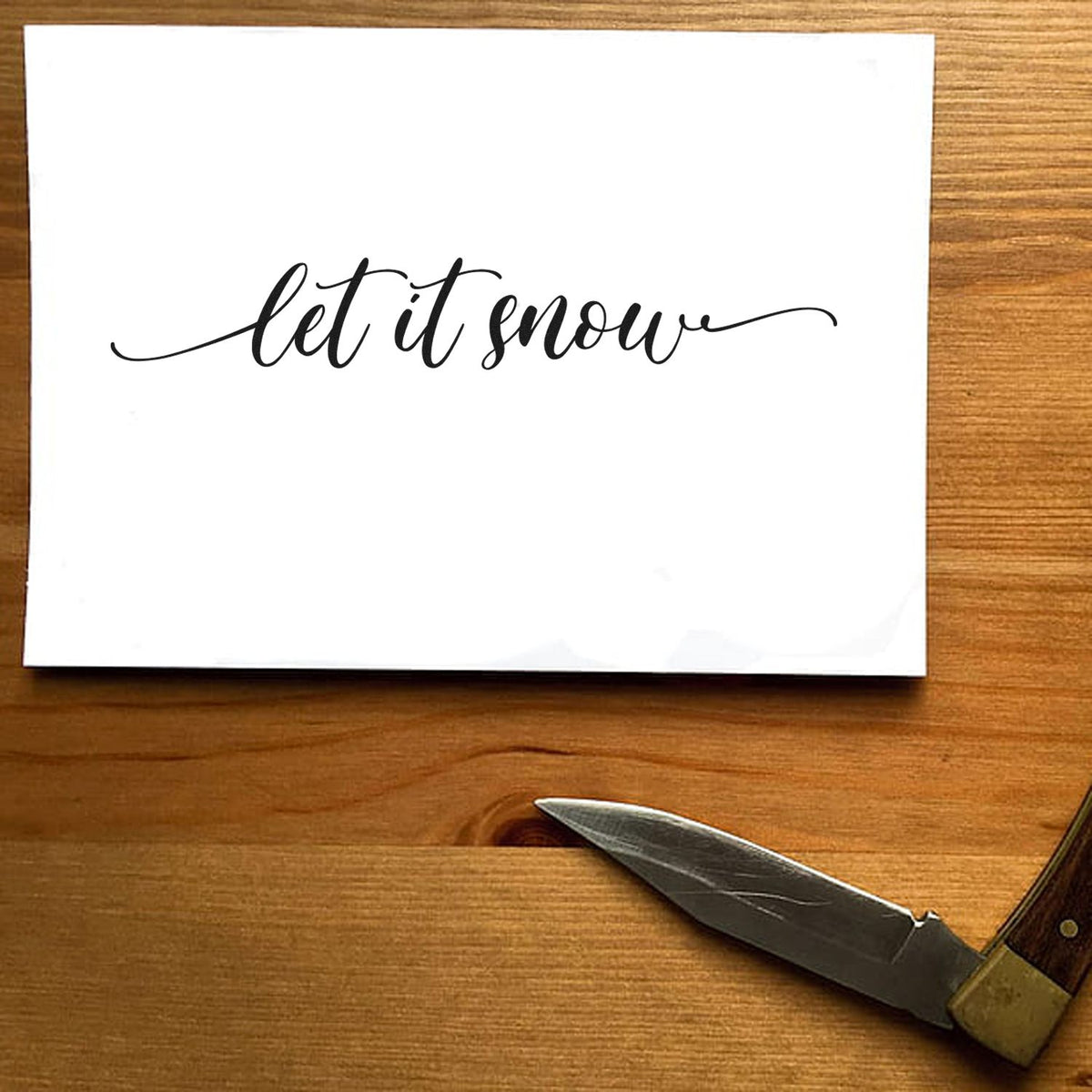 White card with black let it snow script stamp, beside a silver sealing knife on a warm wood table.