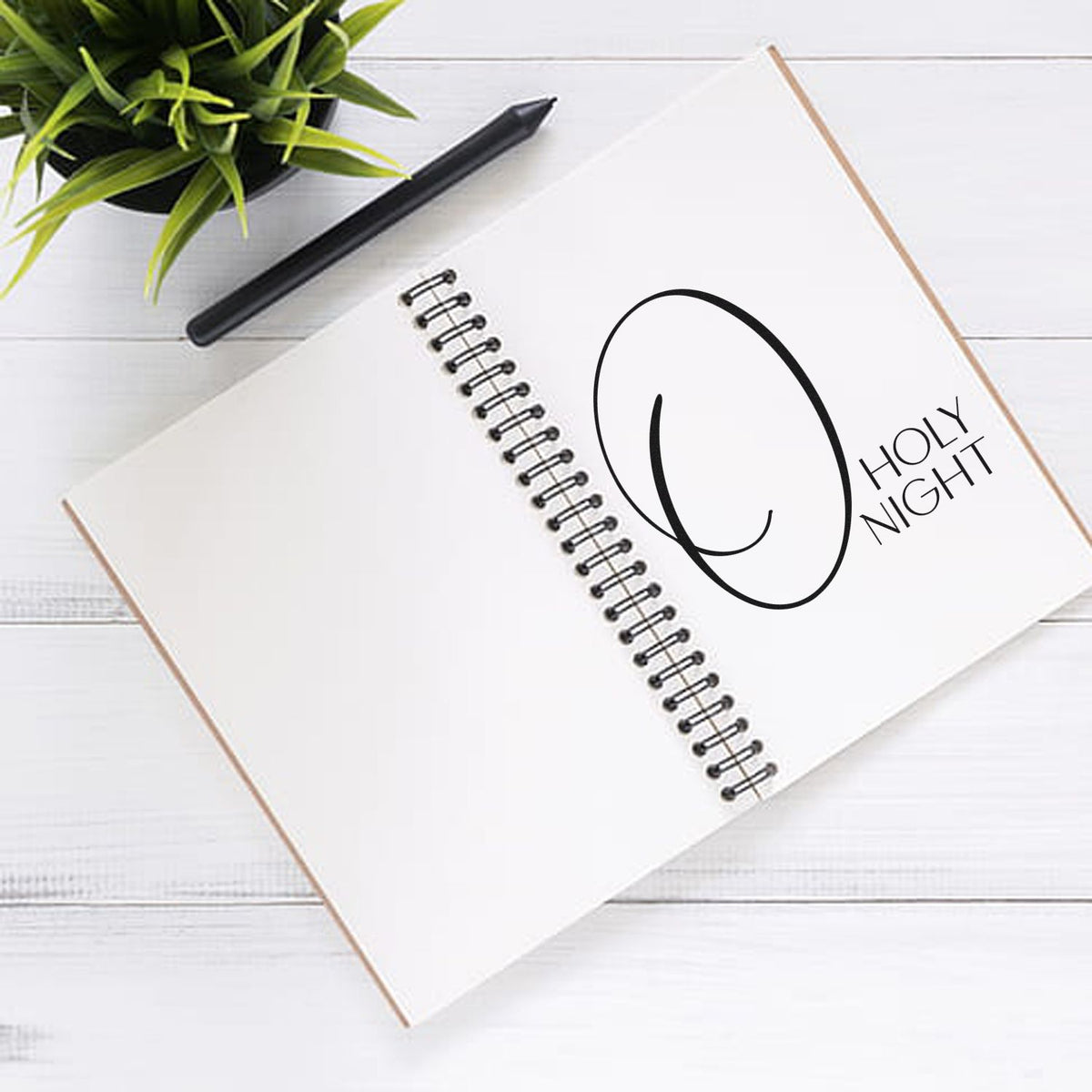White spiral notebook with black Holy Night stamp seal, black pen, and green plant on a white wood backdrop.