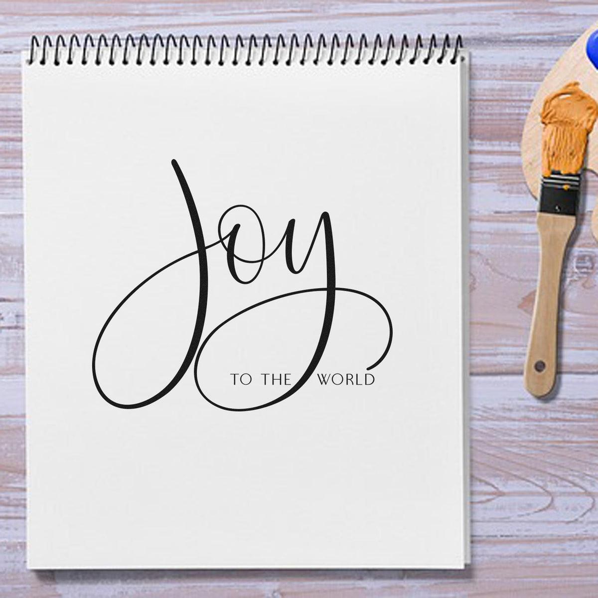 White spiral notebook with black Joy to the World calligraphy, beside a wooden brush and orange wax seal stamp.