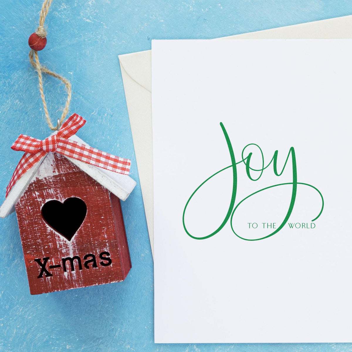 Rustic red birdhouse ornament beside a white holiday card with green Joy to the World stamp and seal on blue background