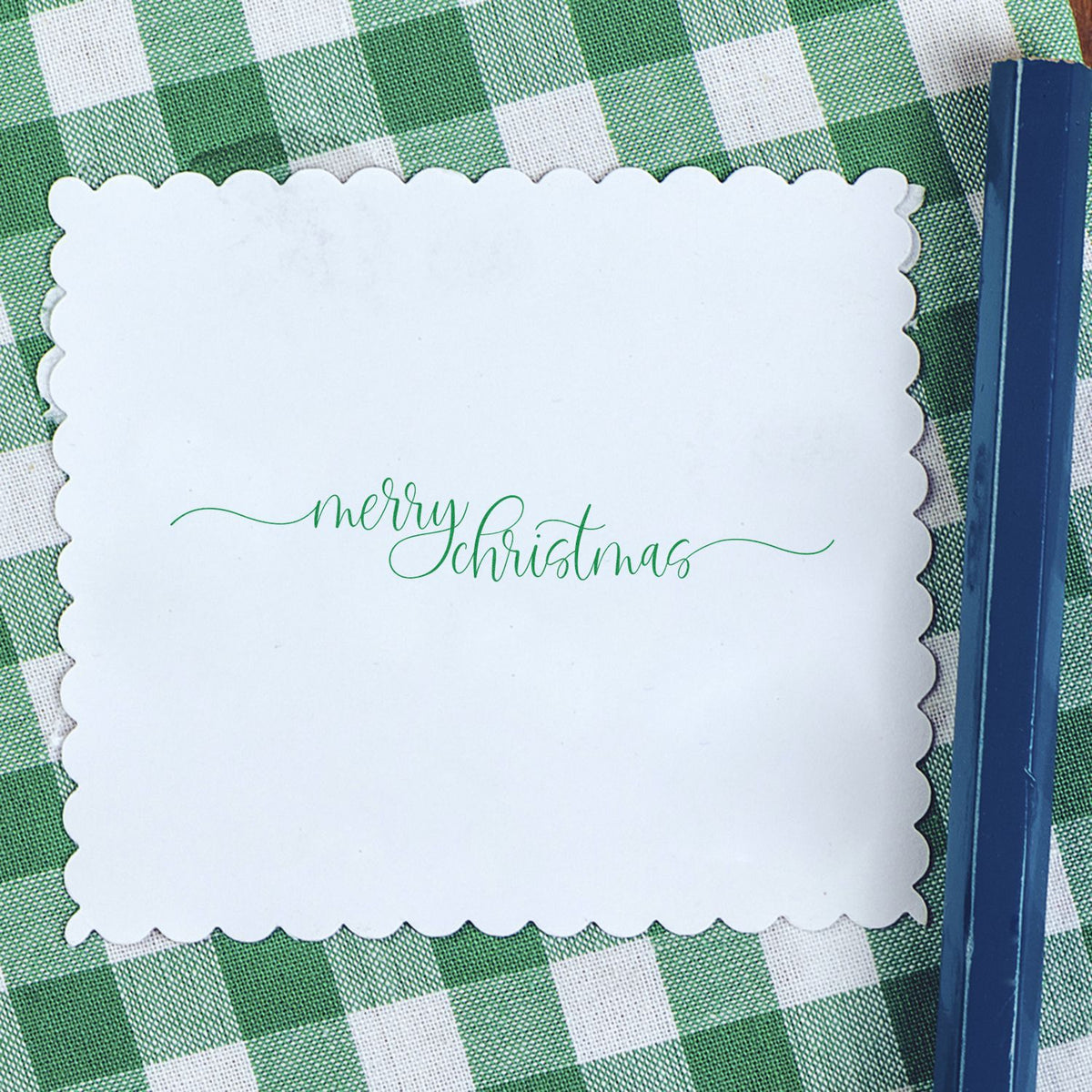 White scalloped Christmas stamp and seal with green merry Christmas script on green gingham, beside a navy blue tool.