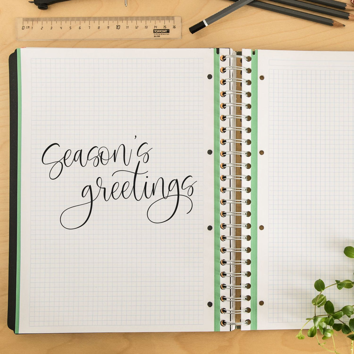 Black calligraphy stamp and seal reading Seasons greetings on graph-paper notebook, with green binding and ruler