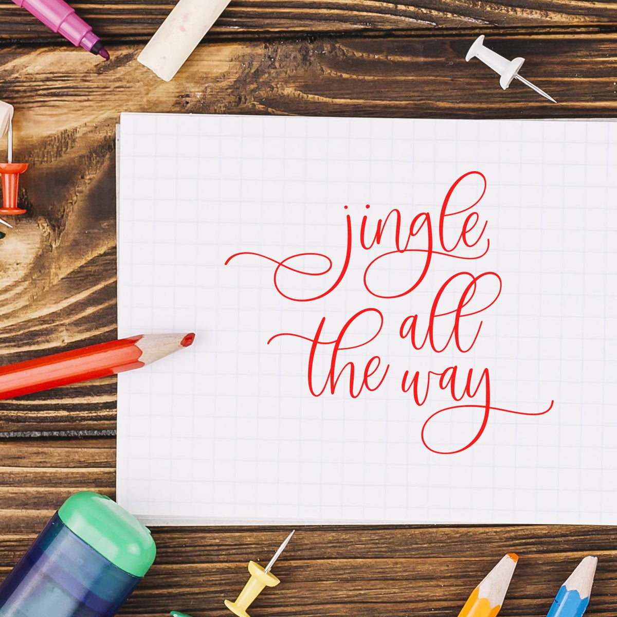 Red calligraphy stamp with jingle all the way on white grid paper, surrounded by pencils and seal tools on rustic wood.