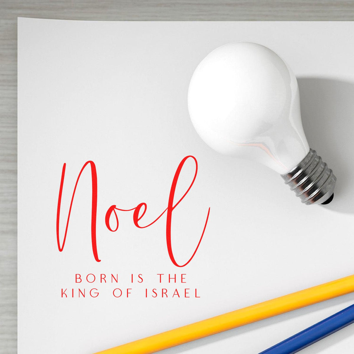 White bulb-shaped stamp seal with red Noel text and Born is the King of Israel on white card, with yellow and blue pencils.