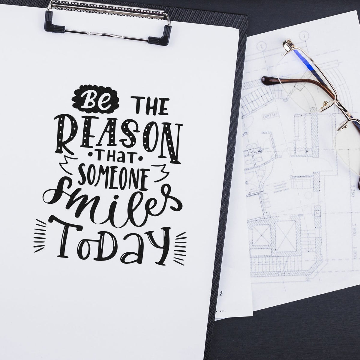 Black stamp and seal design on white paper, reading Be the reason someone smiles today, with pale blue blueprint sheets.