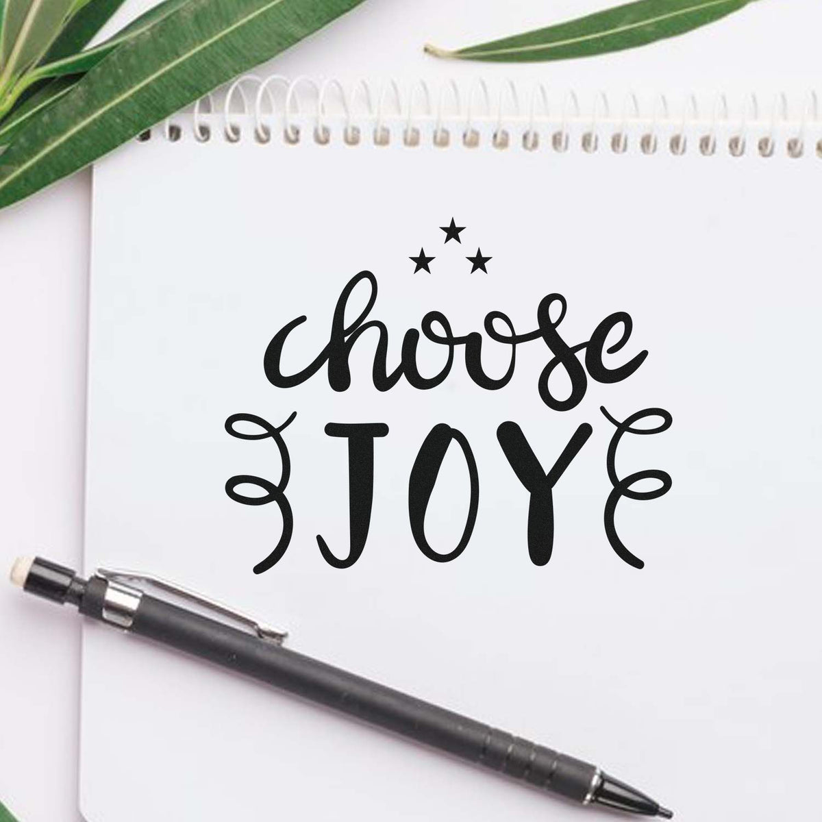 Decorative stamp and seal: black choose JOY text with stars on a white notebook, plus a black pen and green leaves.