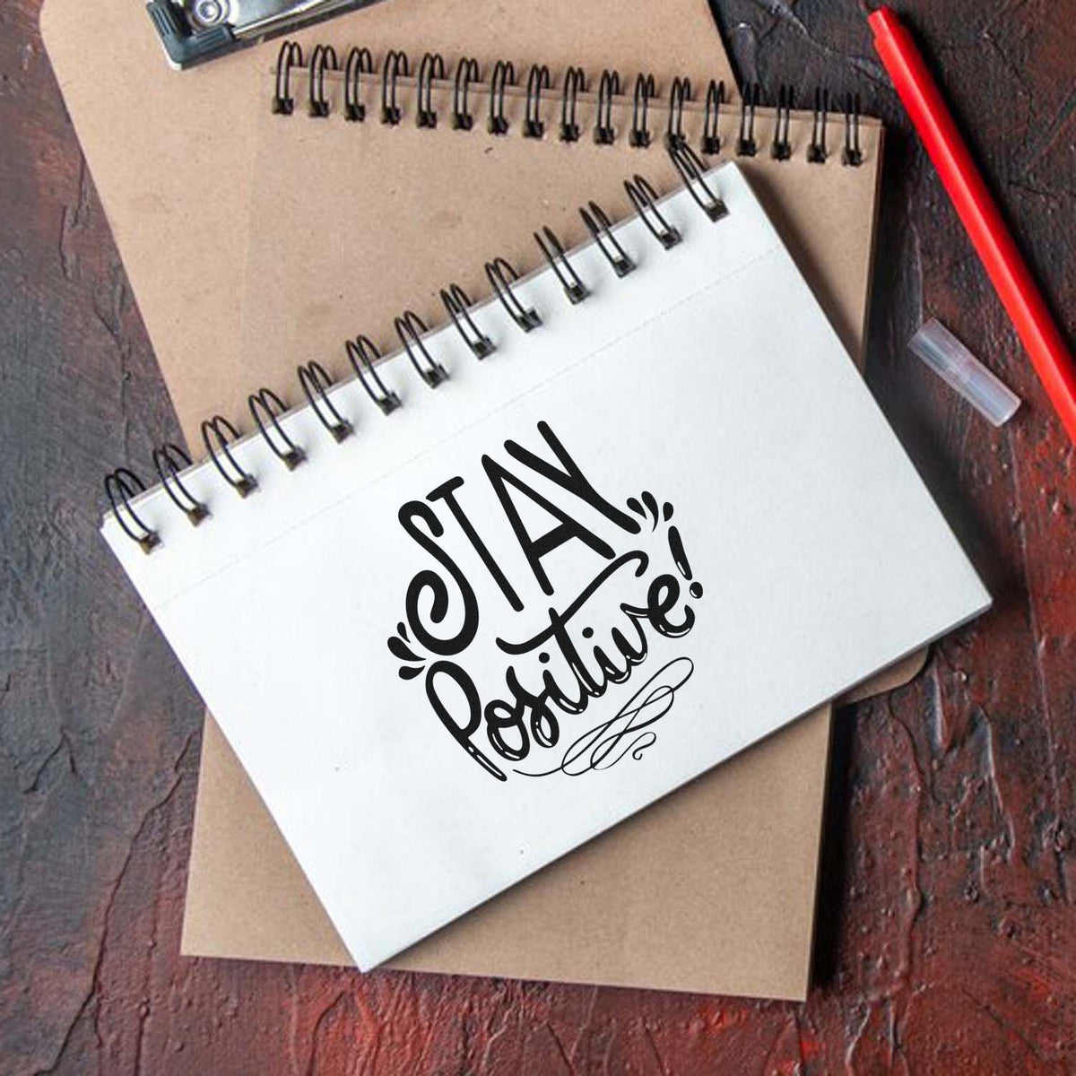 Black Stay Positive! stamp seal on a white spiral notebook over a kraft pad, with a red pen on rustic wood.
