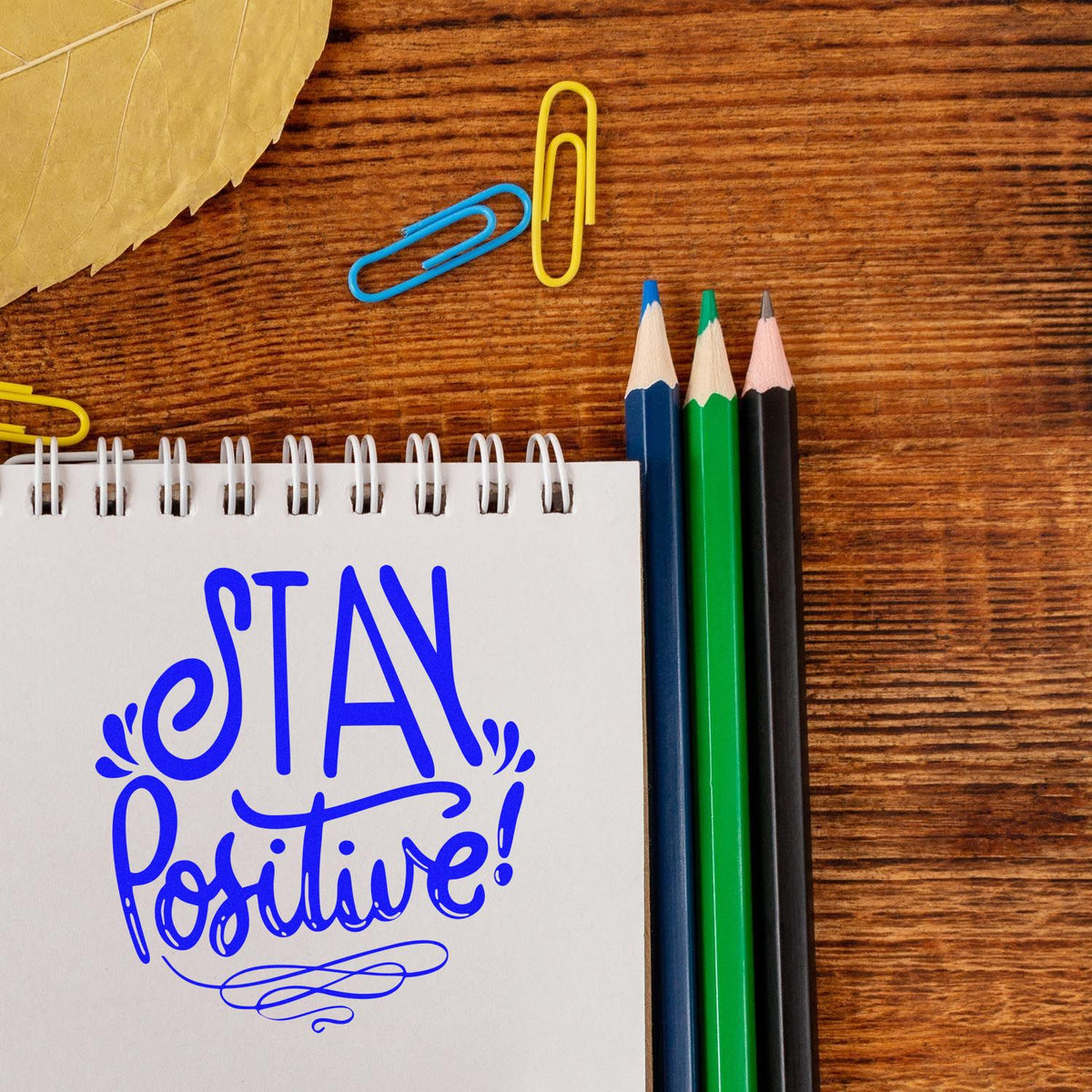 Blue Stay Positive stamp-style seal on a white spiral notebook, with colored pencils and paper clips on a wood desk.