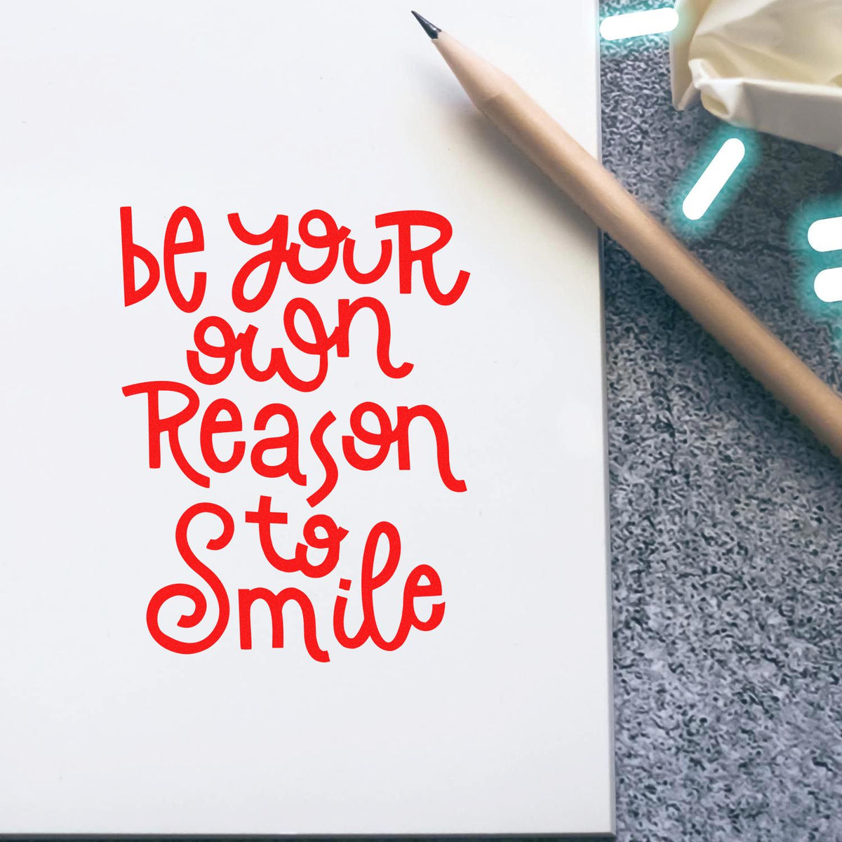 Red stamp and seal design reading be your own reason to smile on white paper, with pencil and grey textured background.