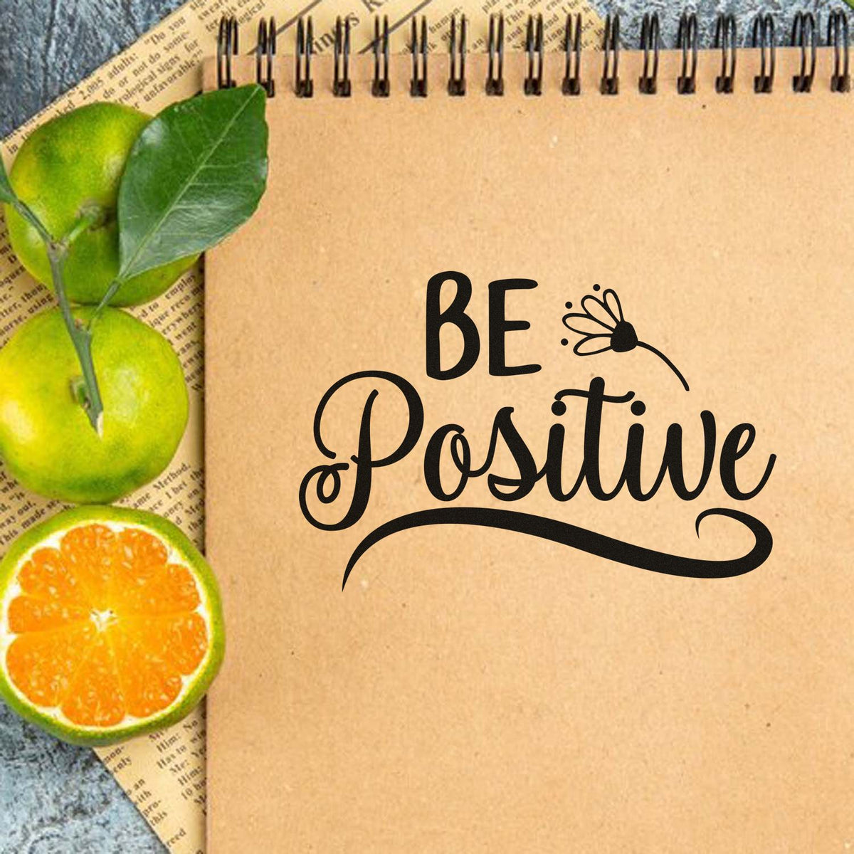 Black BE Positive stamp seal on kraft notebook, with butterfly, green leaves, and sliced green and orange citrus.