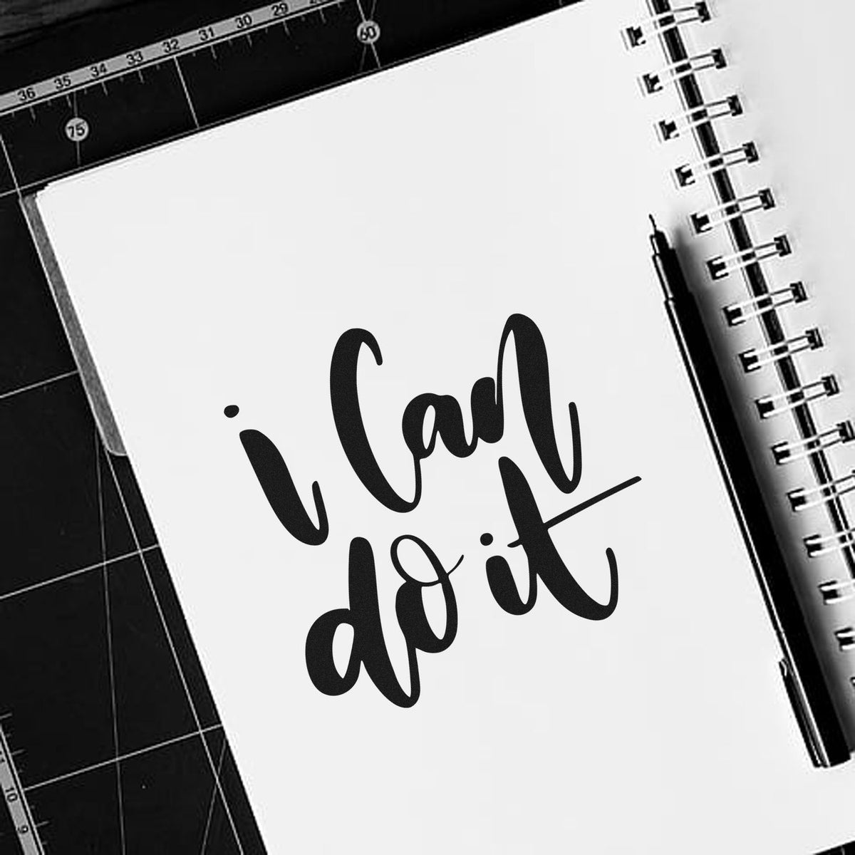 Black stamp and seal design reading I can do it on a white notebook page, with a pen and cutting mat background.