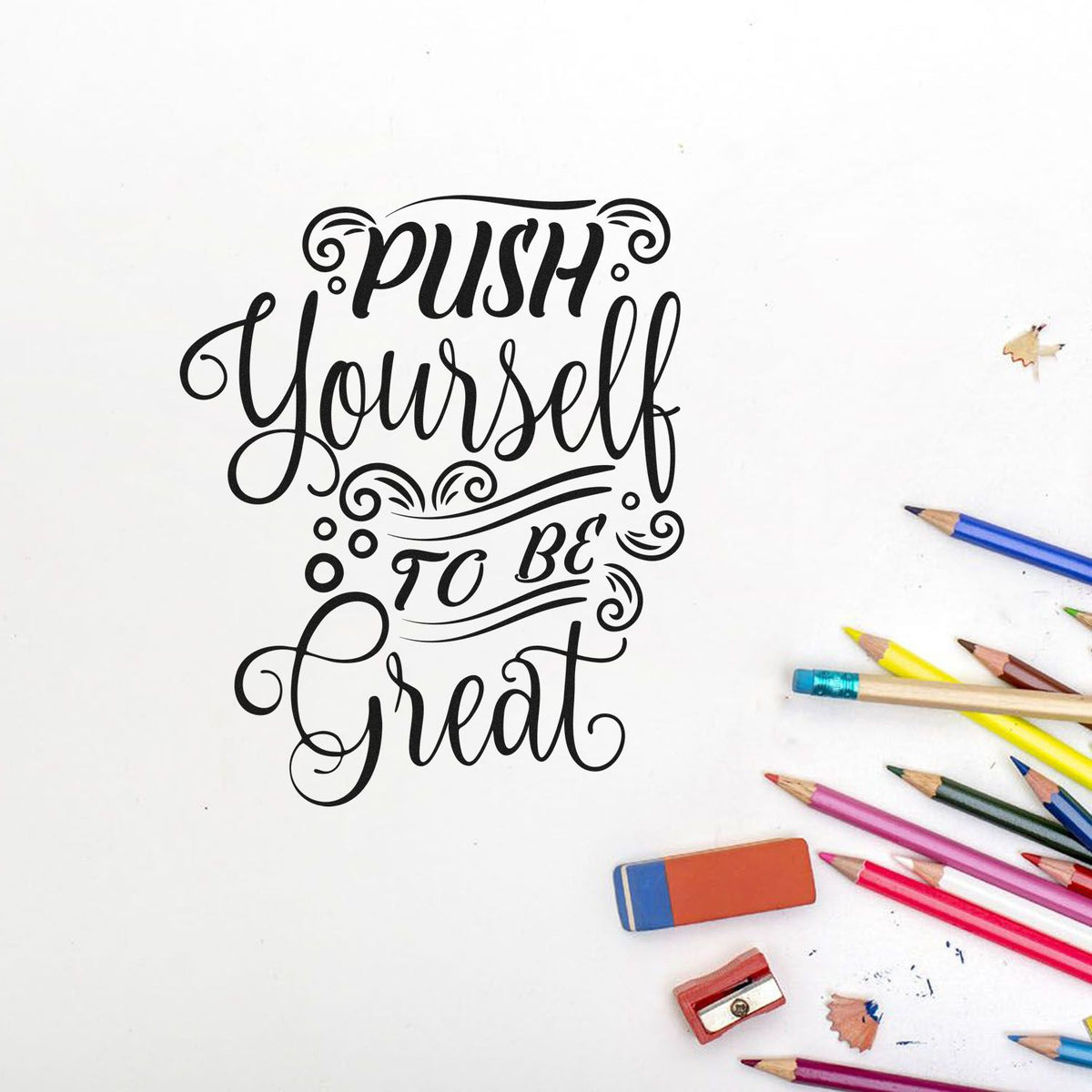 Black decorative stamp and seal reading Push Yourself to Be Great on white, with colorful pencils, eraser, and sharpener.